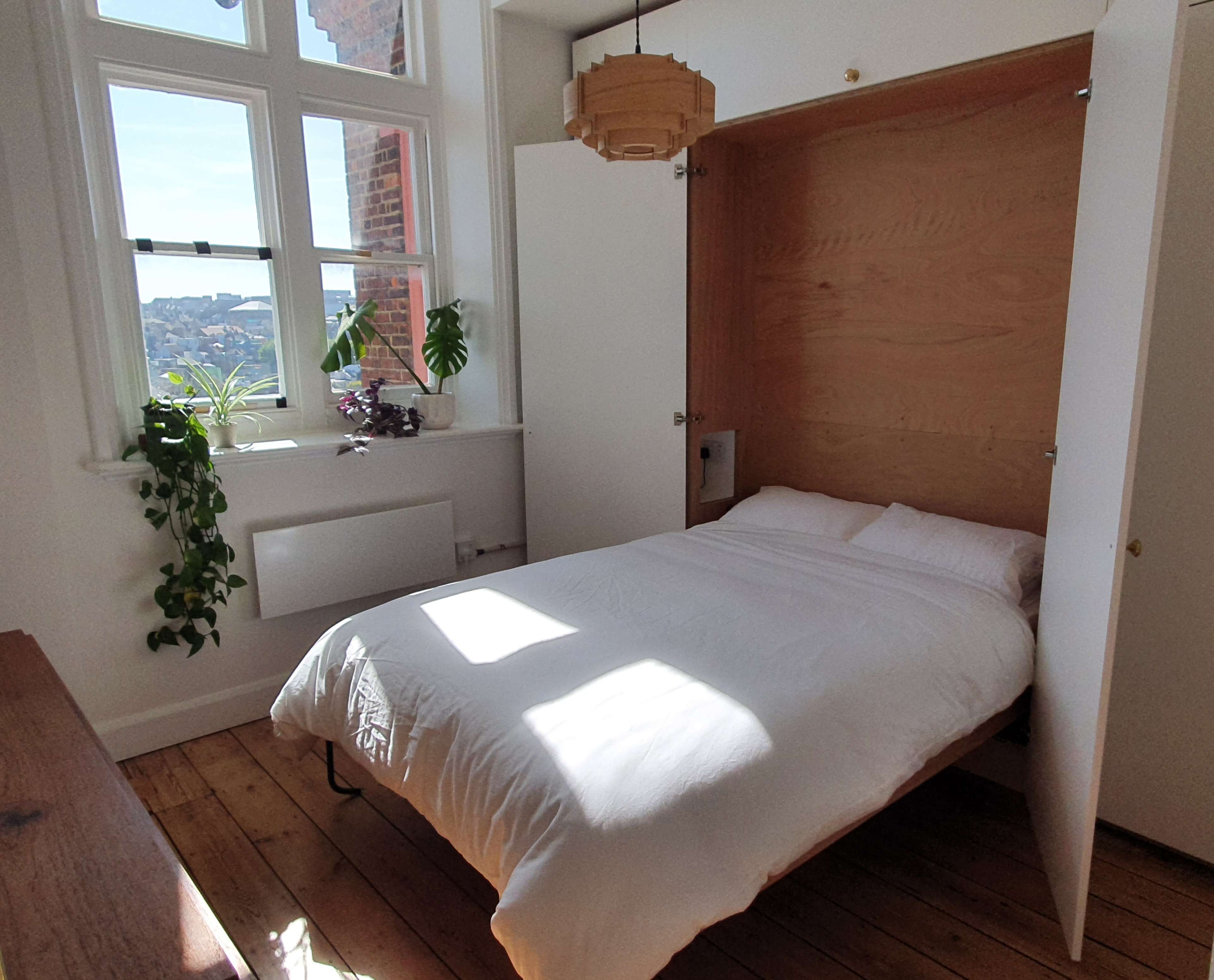 A small bedroom features a fold-down bed with white bedding, sunlight streaming through large windows, and greenery on the walls.