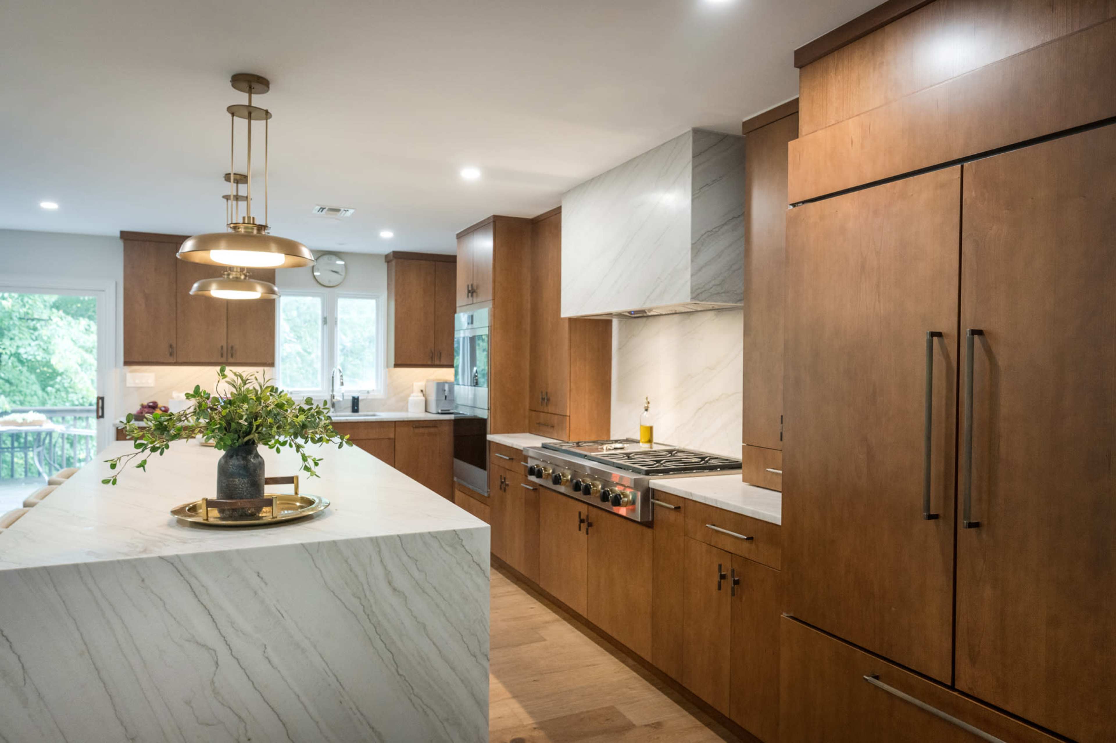 The kitchen features wooden cabinetry, a large marble-topped island, and modern appliances with a view of outdoor greenery through the windows.