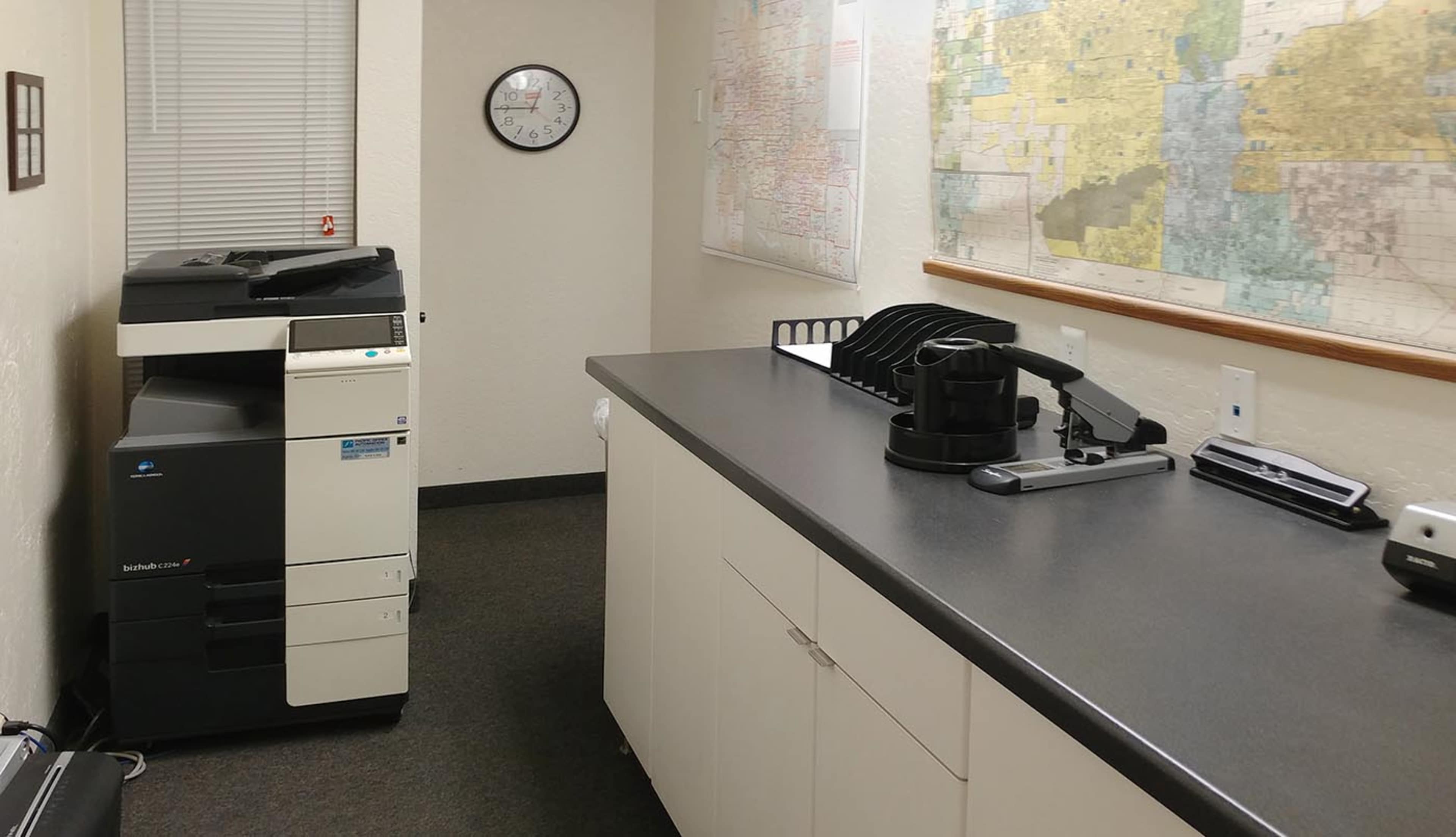 An office space featuring a copier on the left, a clock on the wall, and a large map hanging beside a countertop.