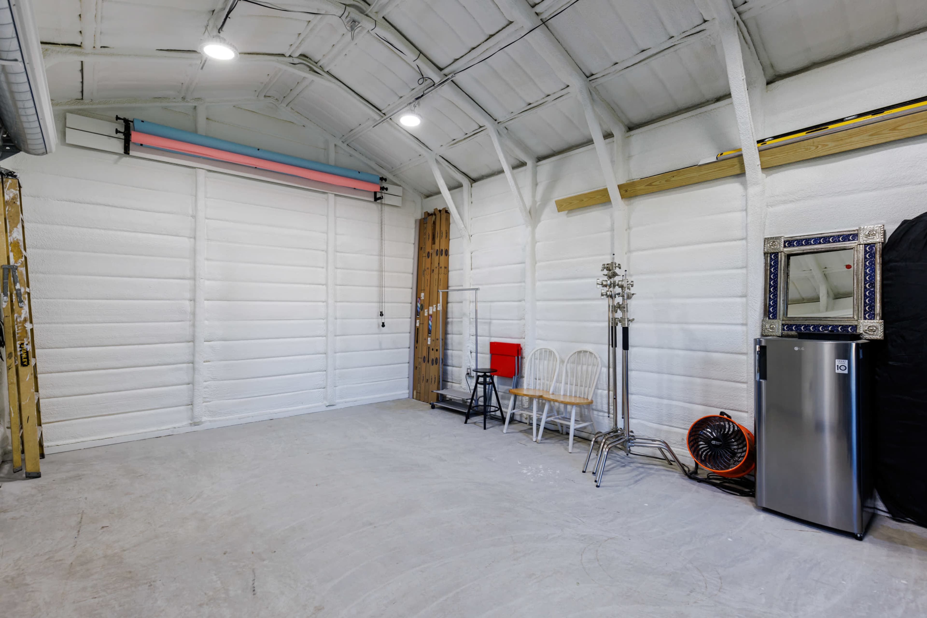 A spacious garage or studio with white insulated walls, a concrete floor, and minimal furnishings, including a red chair, a mirror, and a refrigerator.