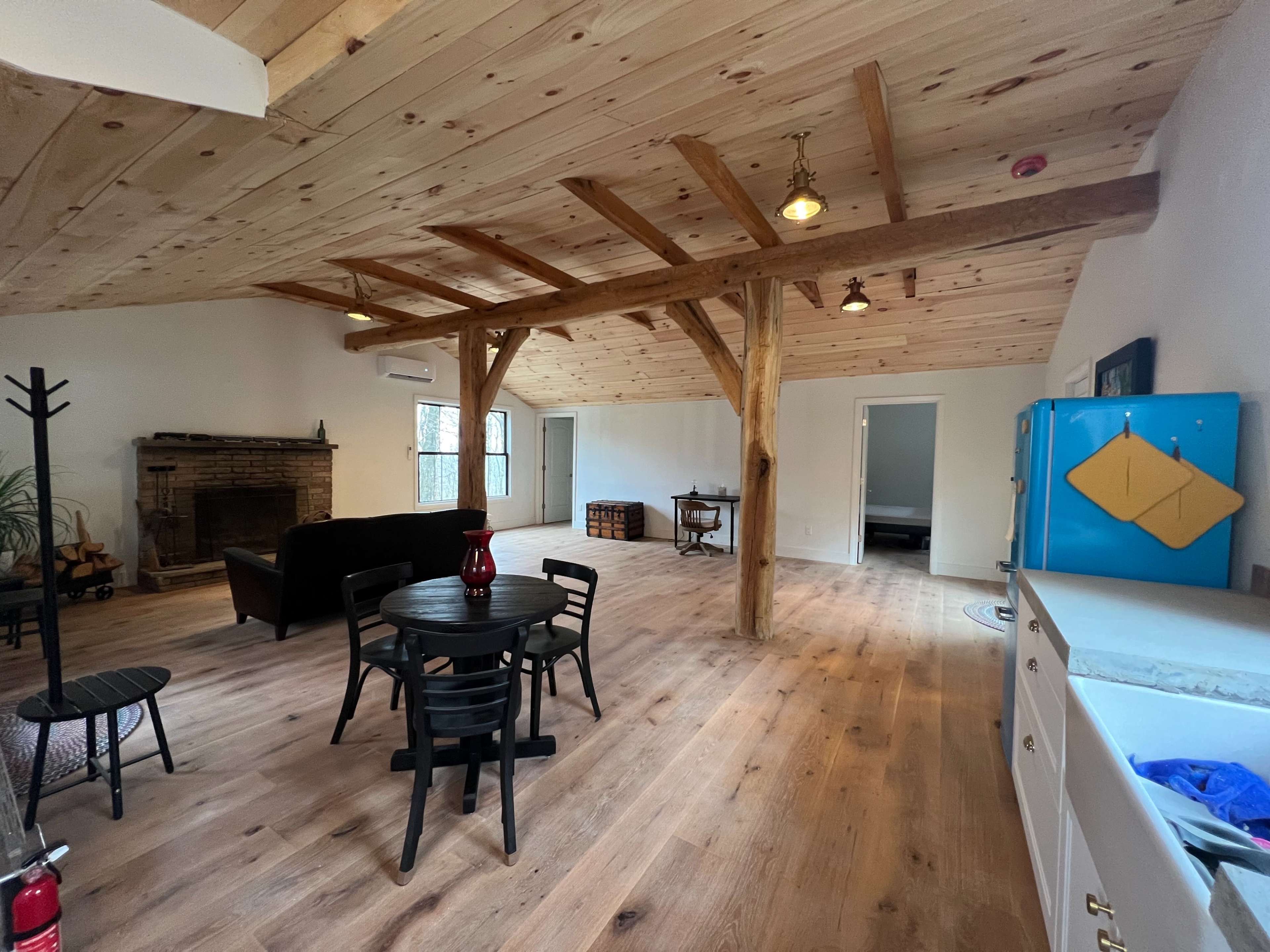A spacious room with wooden beams, a round table and chairs, a blue refrigerator, a fireplace, and several doors leading to other areas.