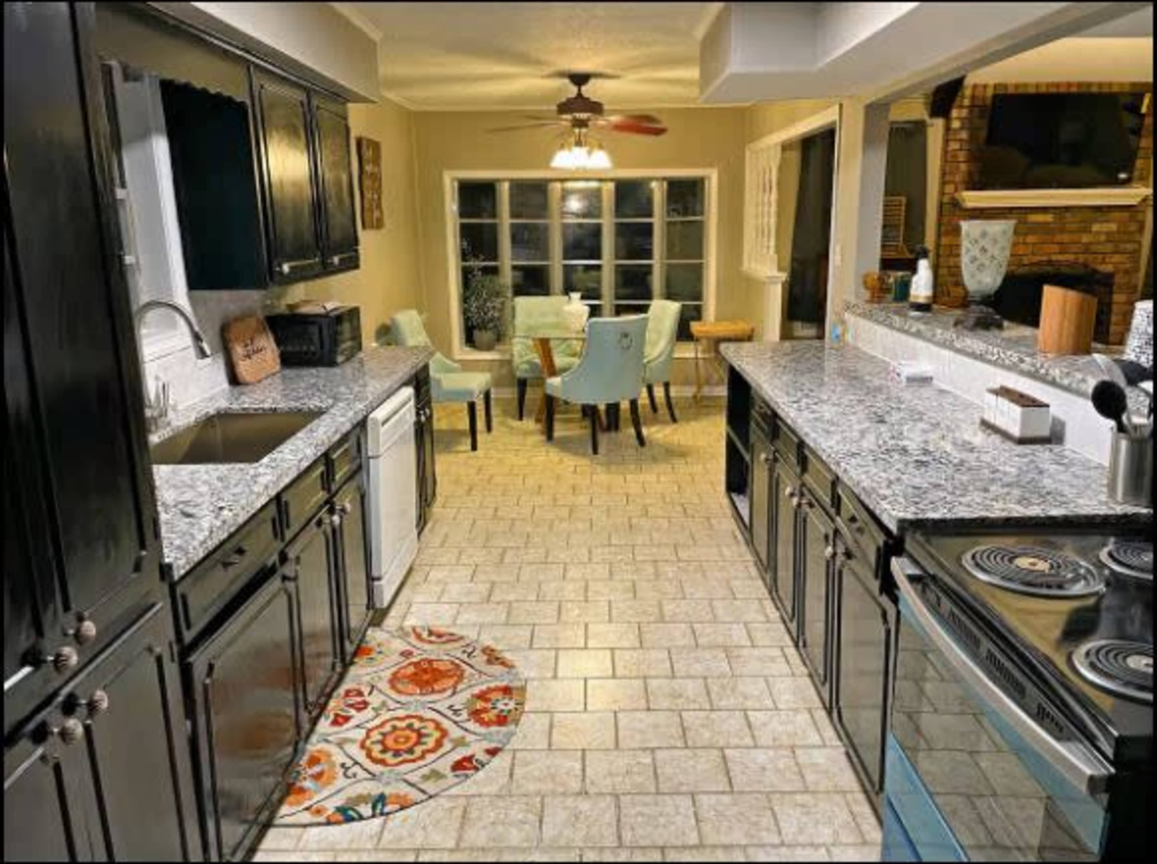 A modern kitchen features dark cabinetry, granite countertops, and a dining area with light-colored chairs, extending into a well-lit space.