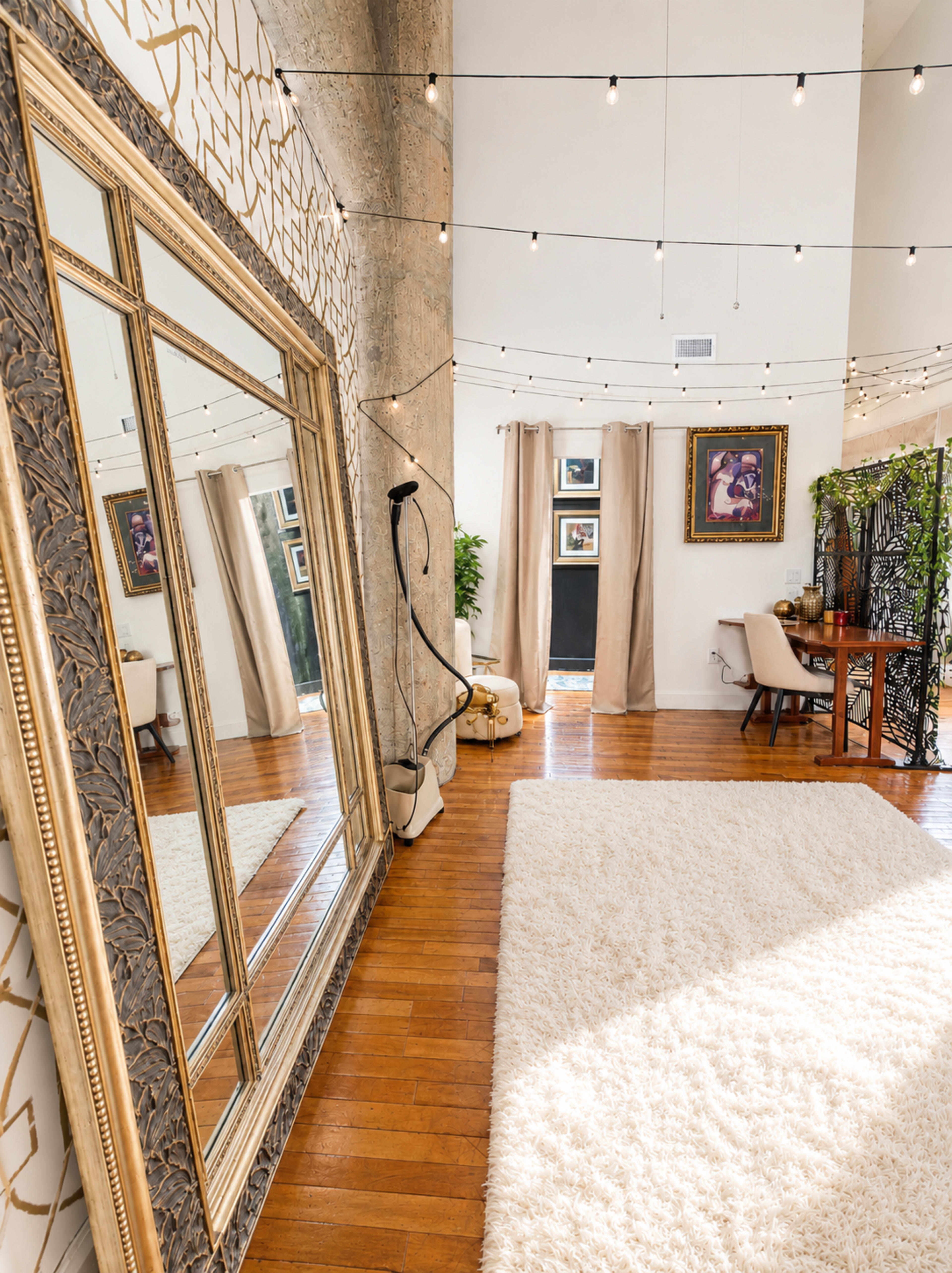 The image depicts a spacious room featuring a large ornate mirror, decorative string lights overhead, and a cozy seating area with a small desk and artwork on the walls.