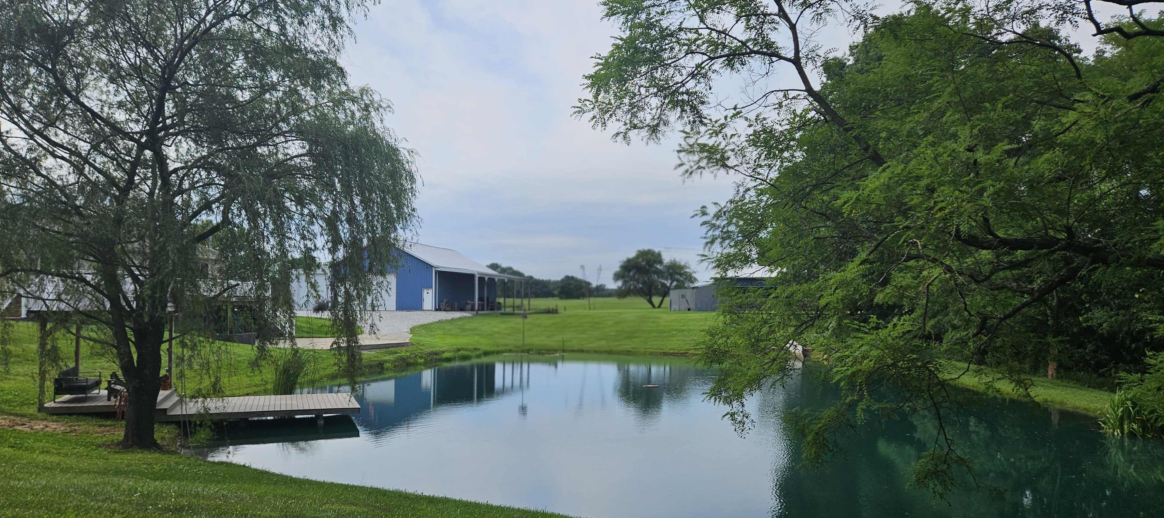 The image shows a serene landscape featuring a pond surrounded by lush greenery, with a blue barn and another structure in the background.