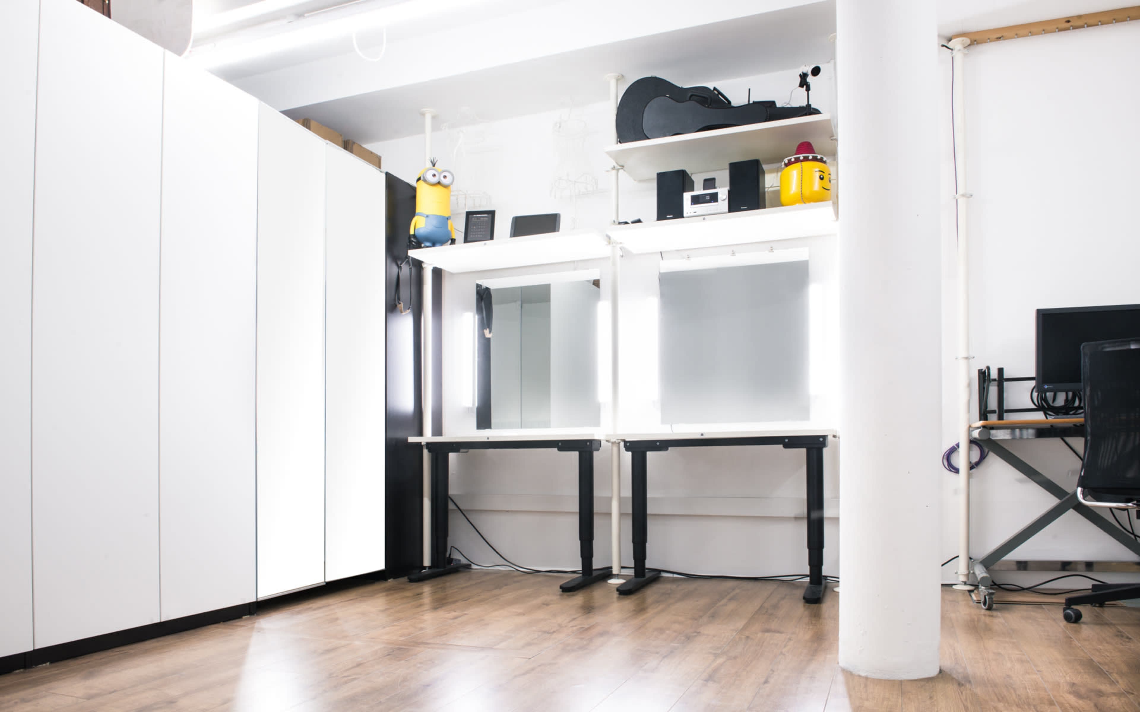A modern workspace with white storage cabinets, adjustable workstations, and various items including a guitar and a character figure on shelves.