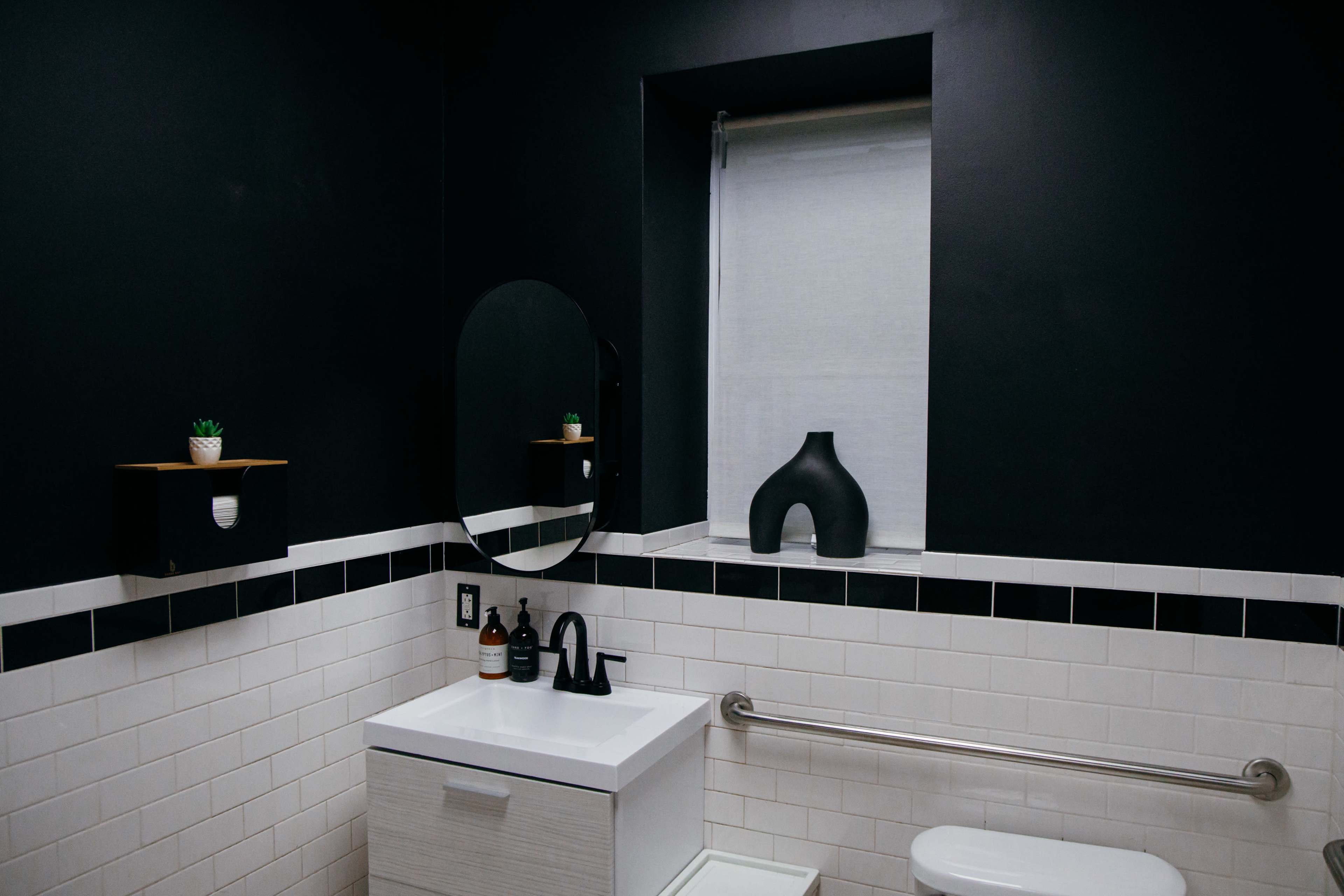 The image shows a modern bathroom with black walls, a white sink cabinet, a round mirror, and a small black vase on the windowsill.