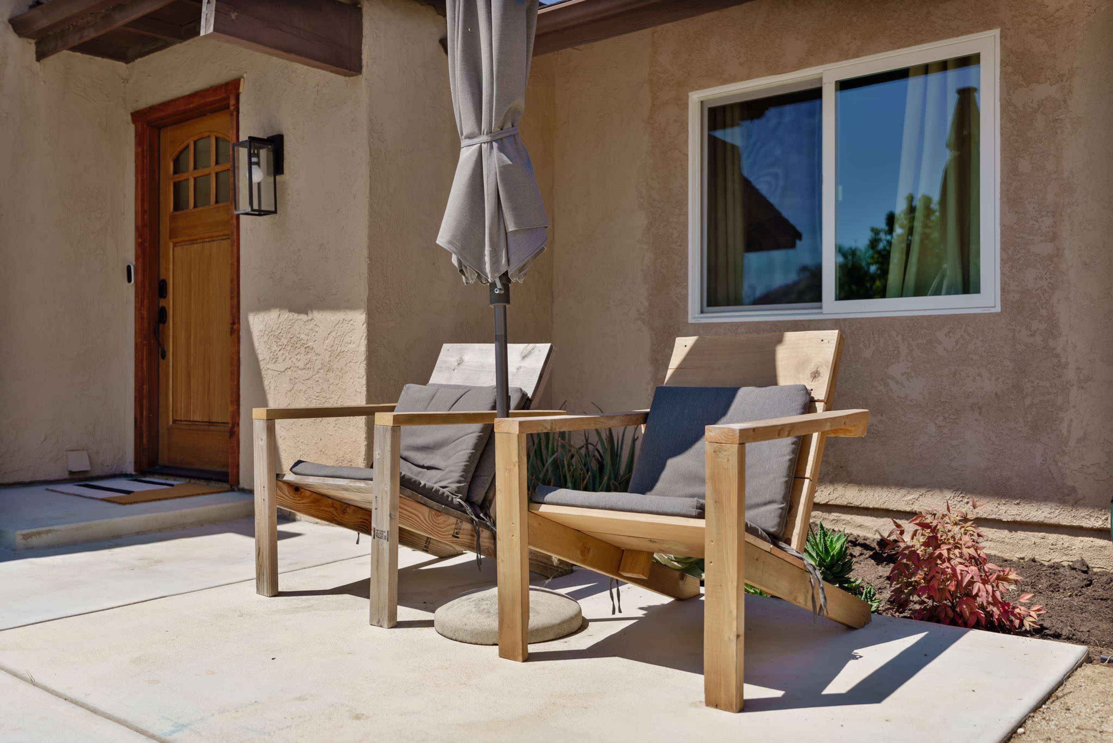 Two wooden lounge chairs with gray cushions and an umbrella stand beside a house entryway.