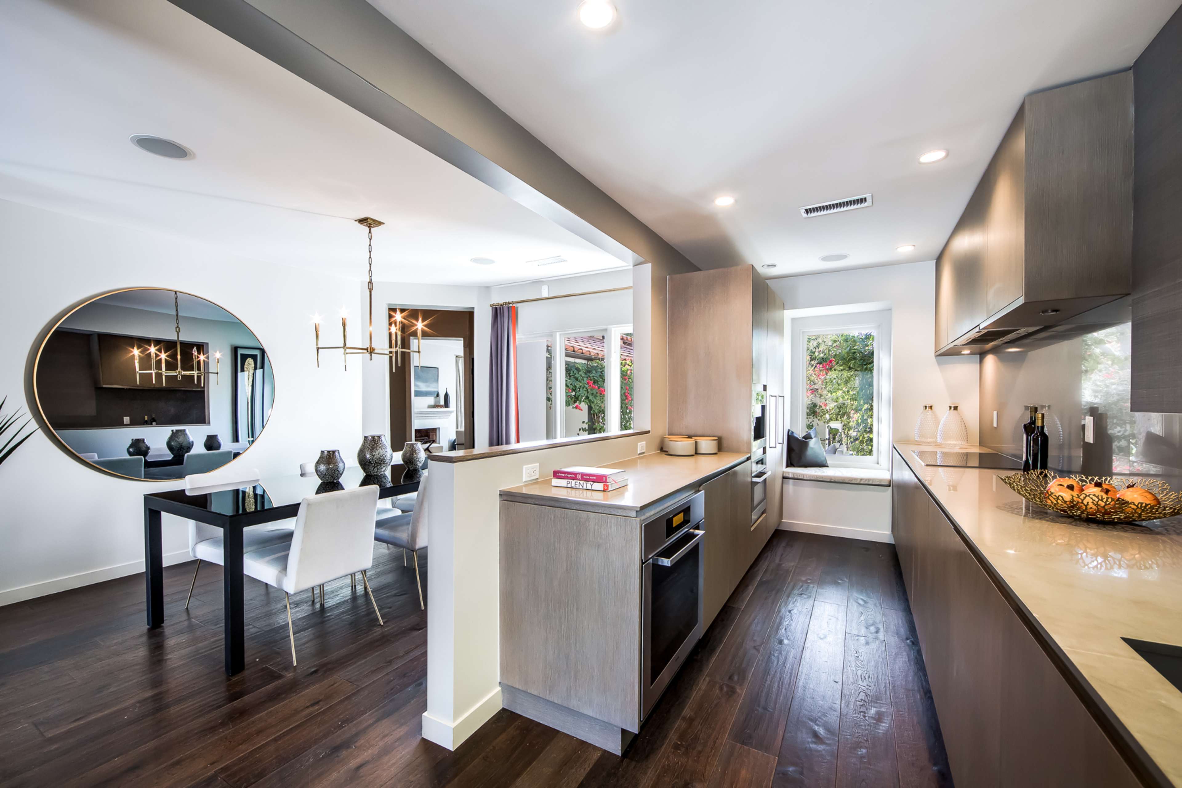 The image shows a modern kitchen with sleek cabinetry, an island, and a dining area featuring a round mirror and a table with chairs.