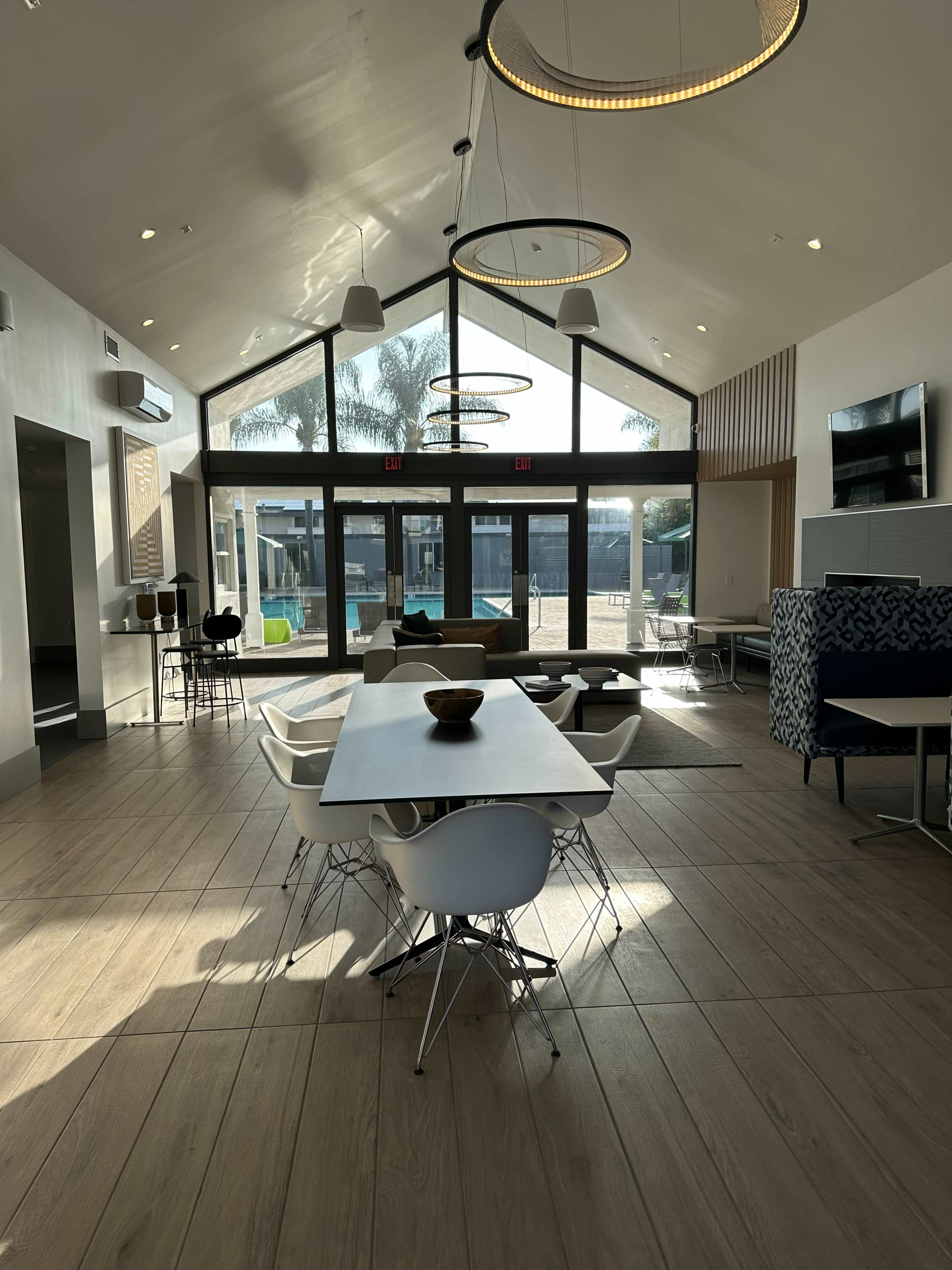 A modern lounge area with a large table and several chairs, featuring large windows that overlook a swimming pool area.