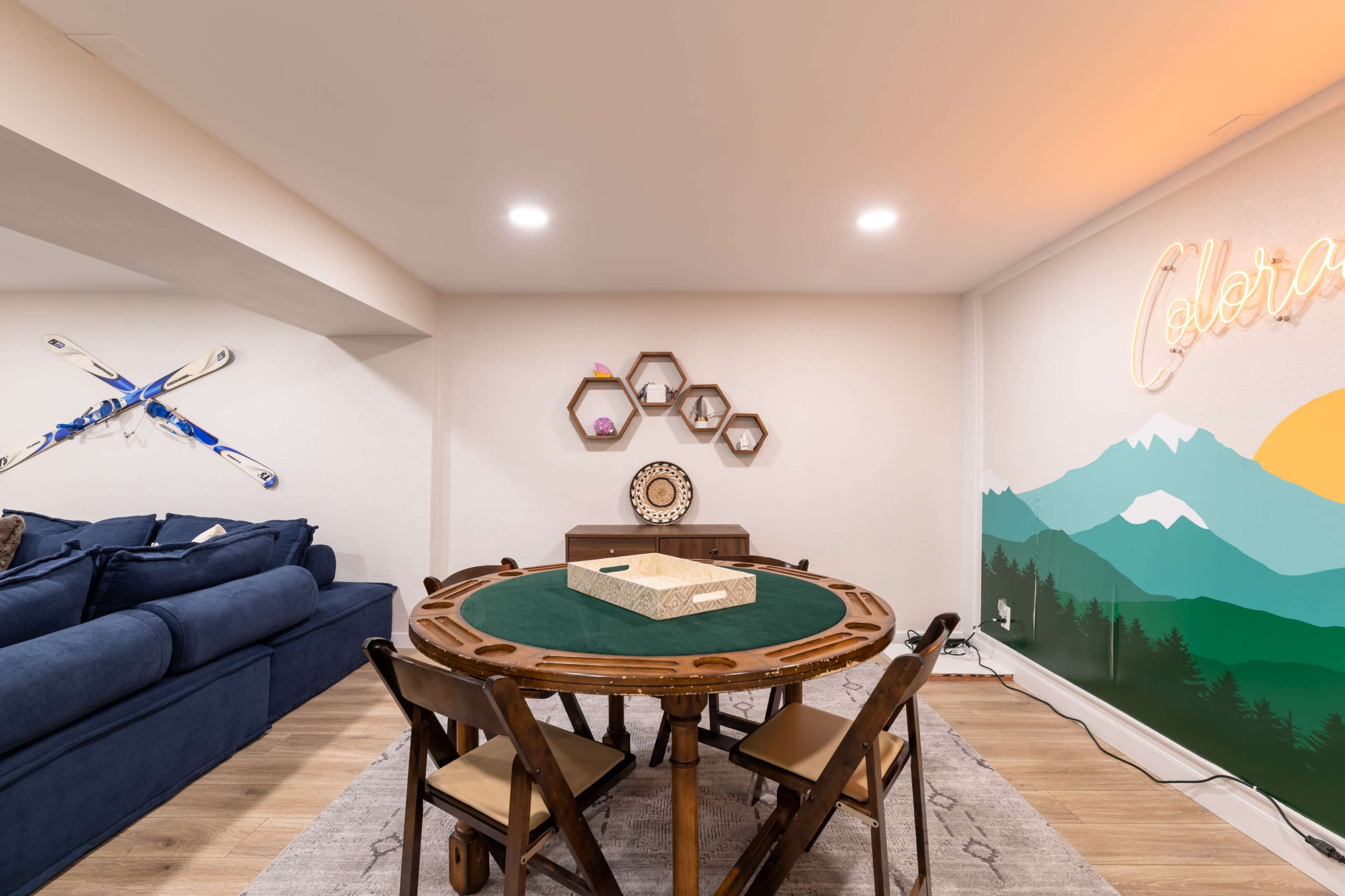 A cozy basement game area featuring a round table with a green felt surface, surrounded by wooden chairs, and a decorated wall with mountain mural art and a neon sign.