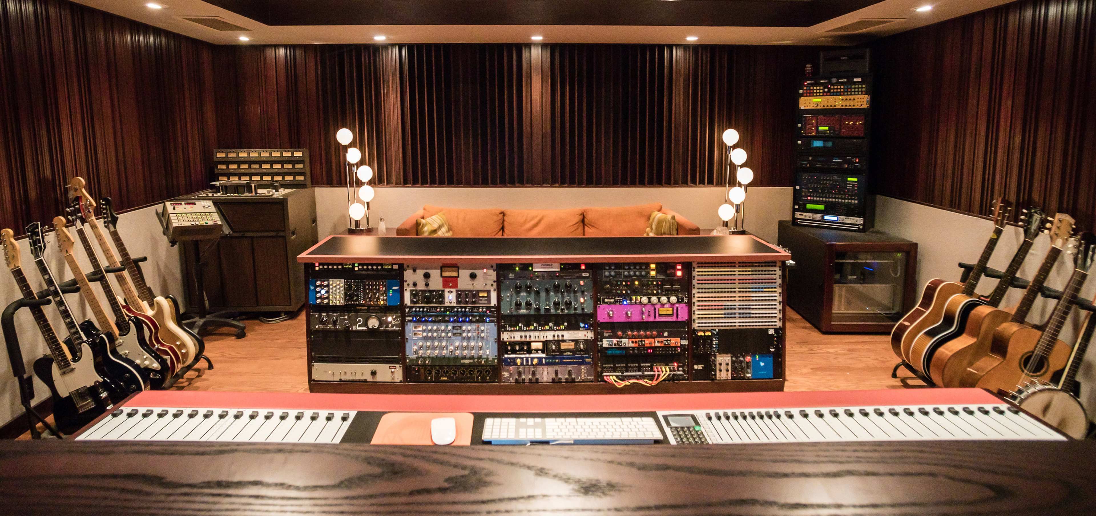 The image shows a music studio featuring an array of audio equipment, a central mixing console, and multiple guitars arranged along the walls.
