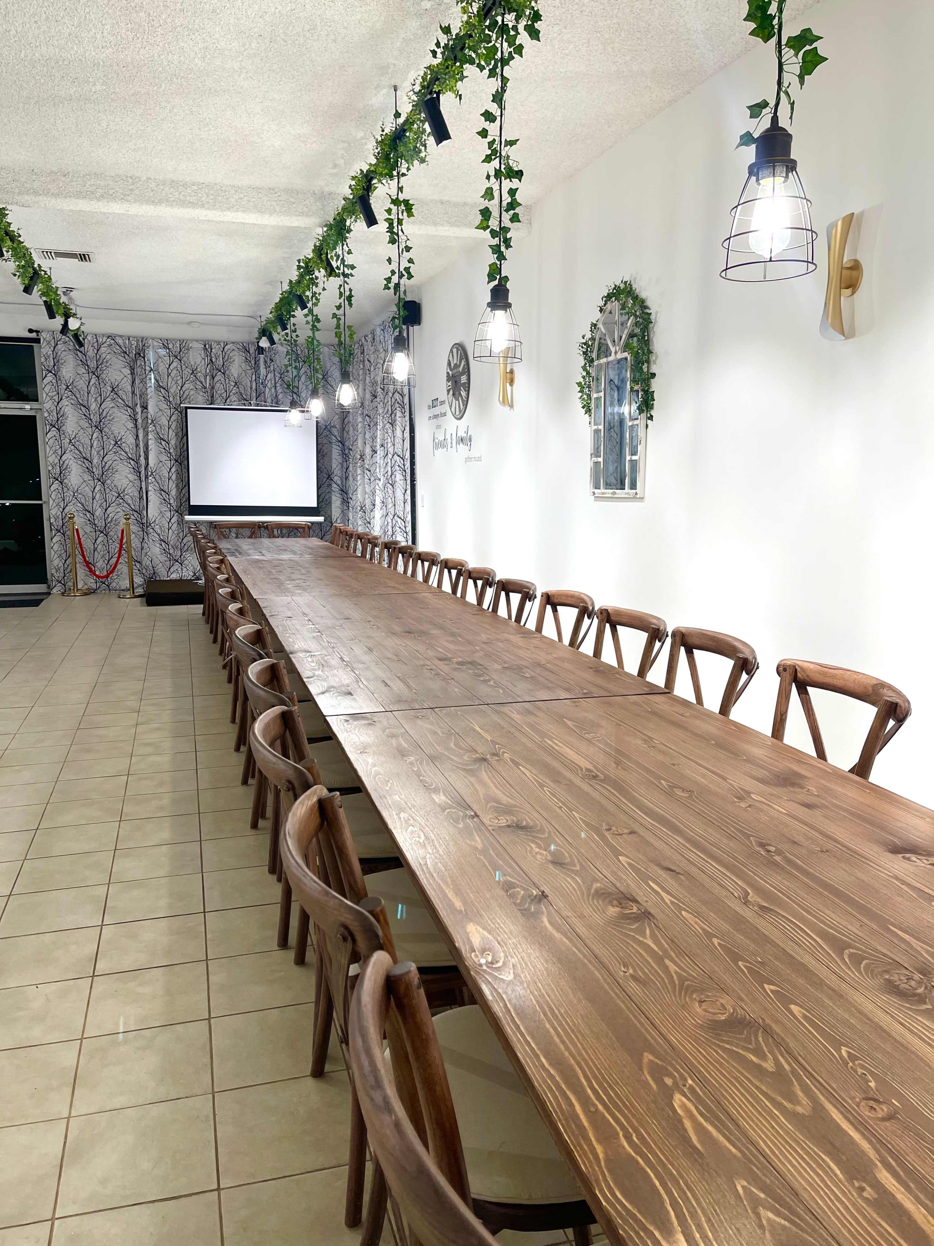 A long wooden table surrounded by chairs in a well-lit room featuring decorative plants and a presentation screen.