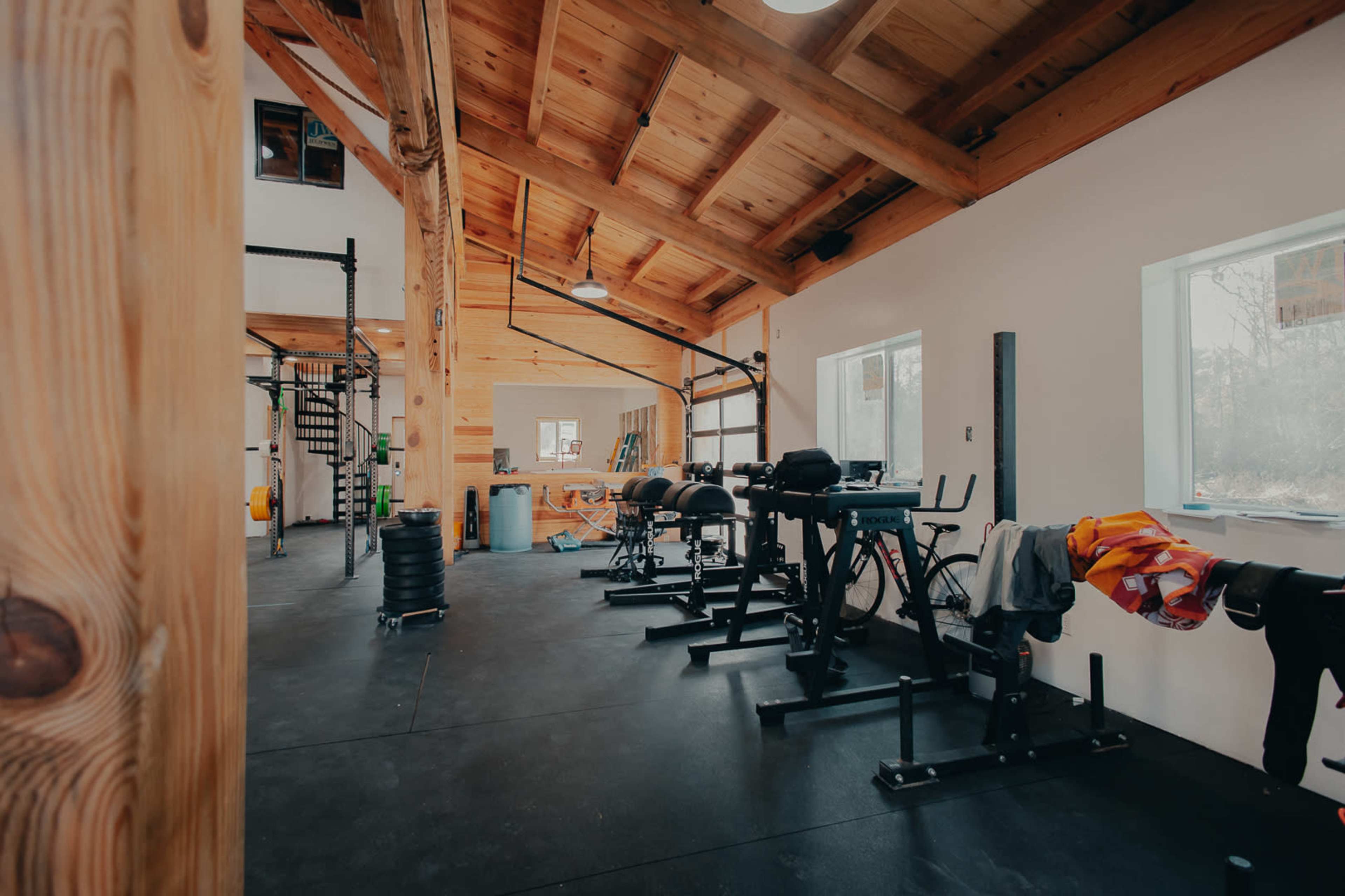 The image shows an interior view of a gym with wooden beams, fitness equipment, and large windows.