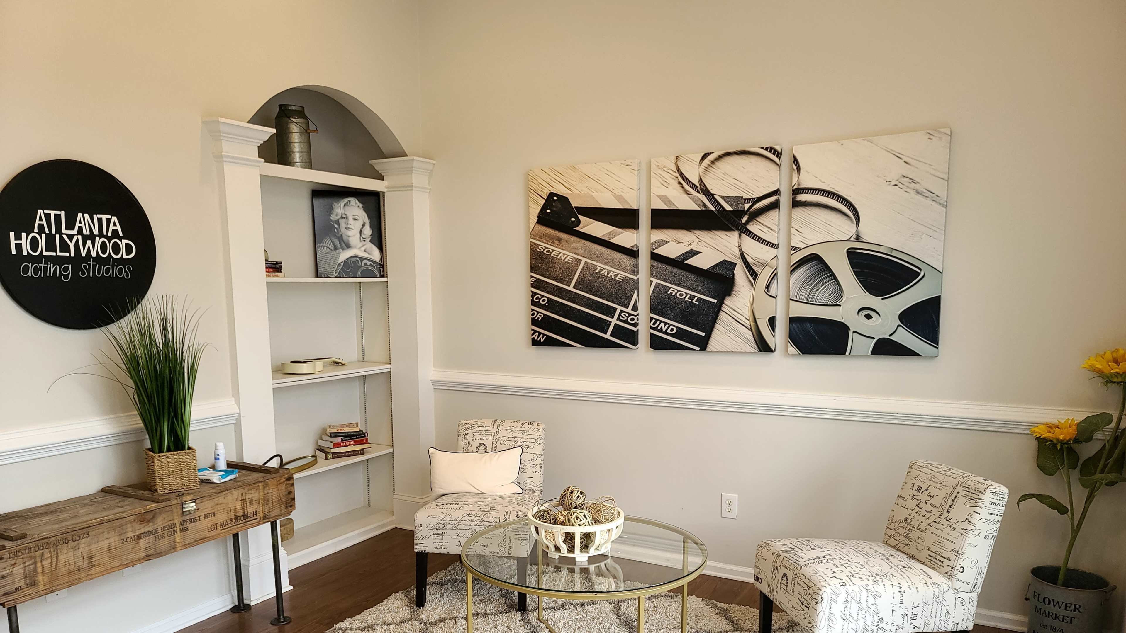 A bright, minimalistic room featuring two patterned chairs, a round glass table, a wooden console, and wall art depicting film-related items.