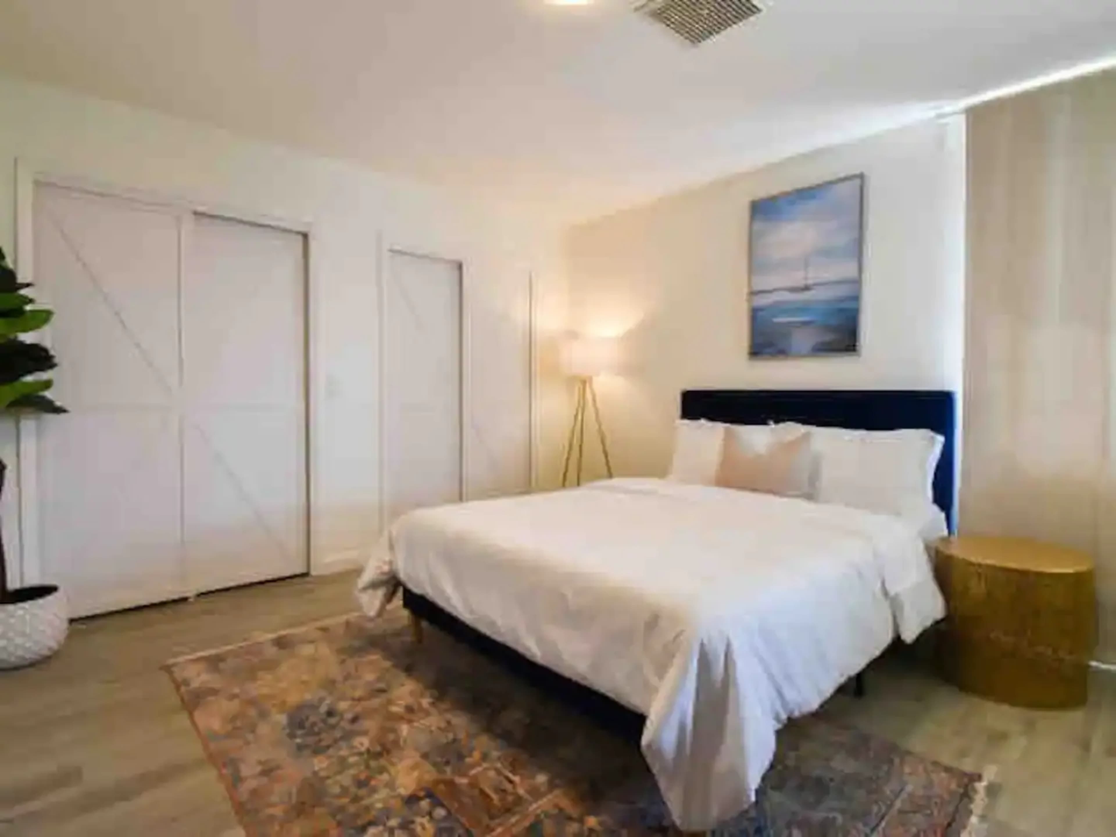 A spacious bedroom features a bed with white linens, a lamp beside it, and a decorative easel with a painting on the wall.
