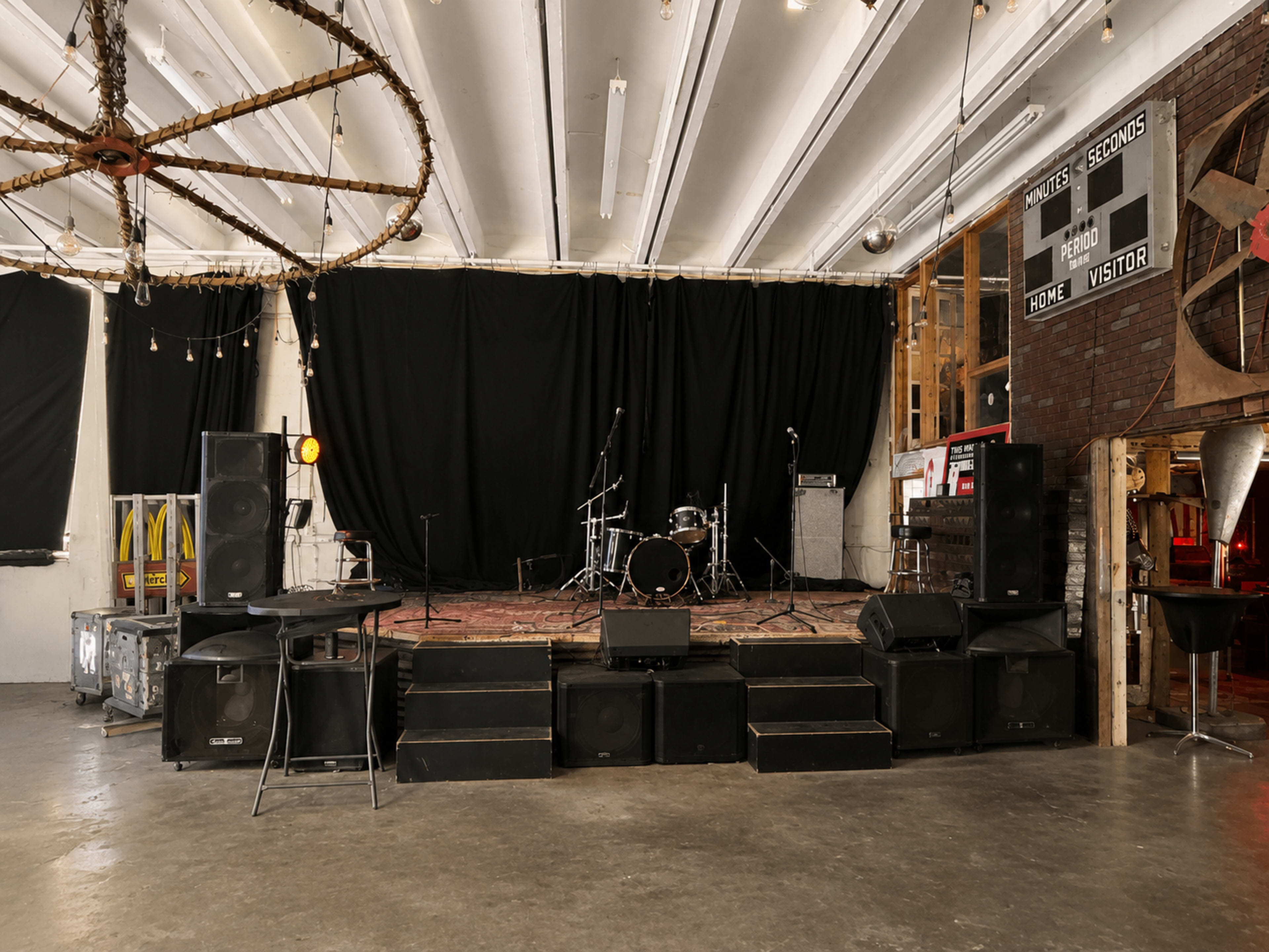 The image shows a performance stage with a drum set, amplifiers, and sound equipment set up in a spacious, industrial-style venue.