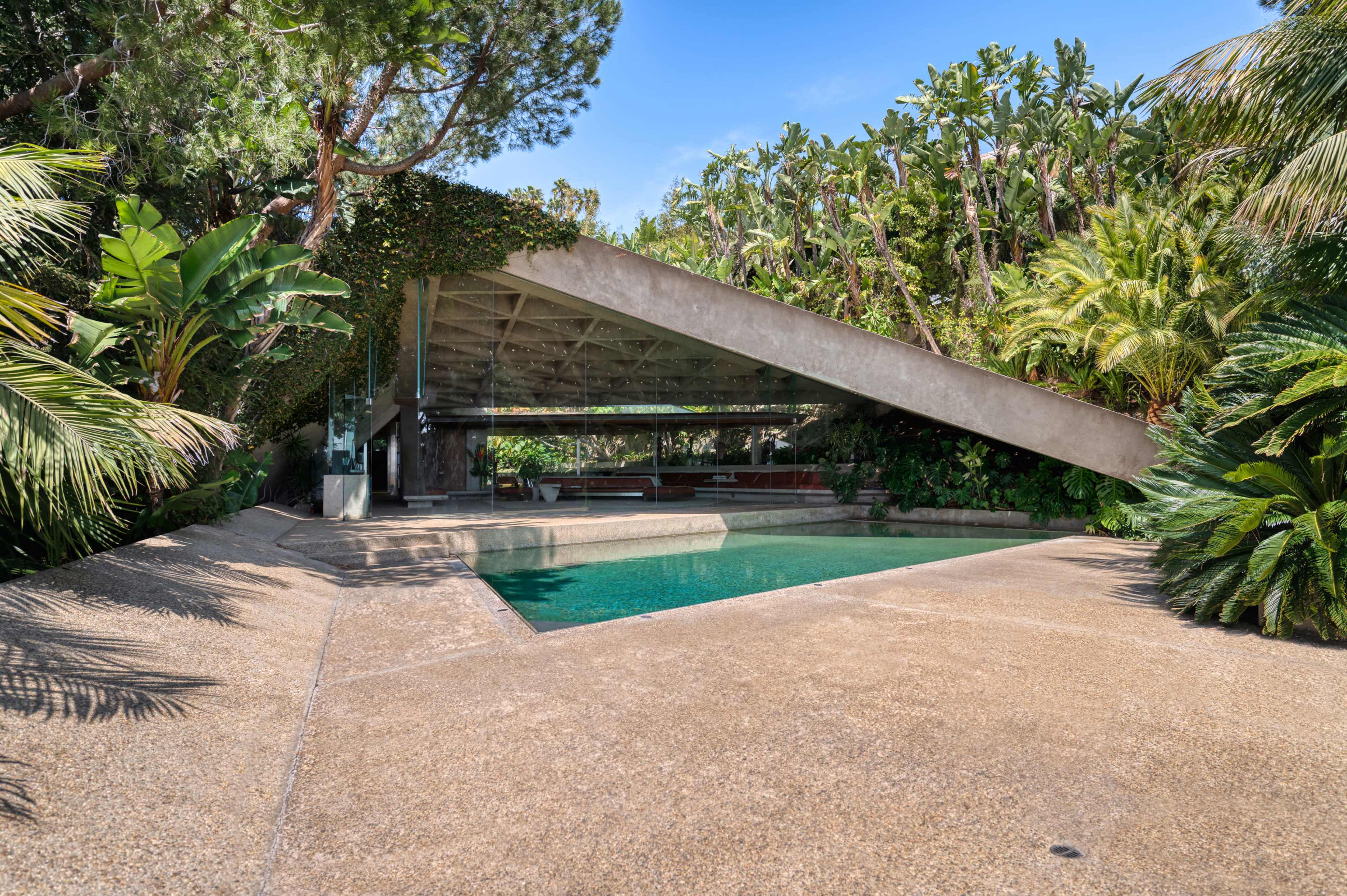 A modern, angular house with a sloping roof is surrounded by lush greenery and features a swimming pool in the foreground.