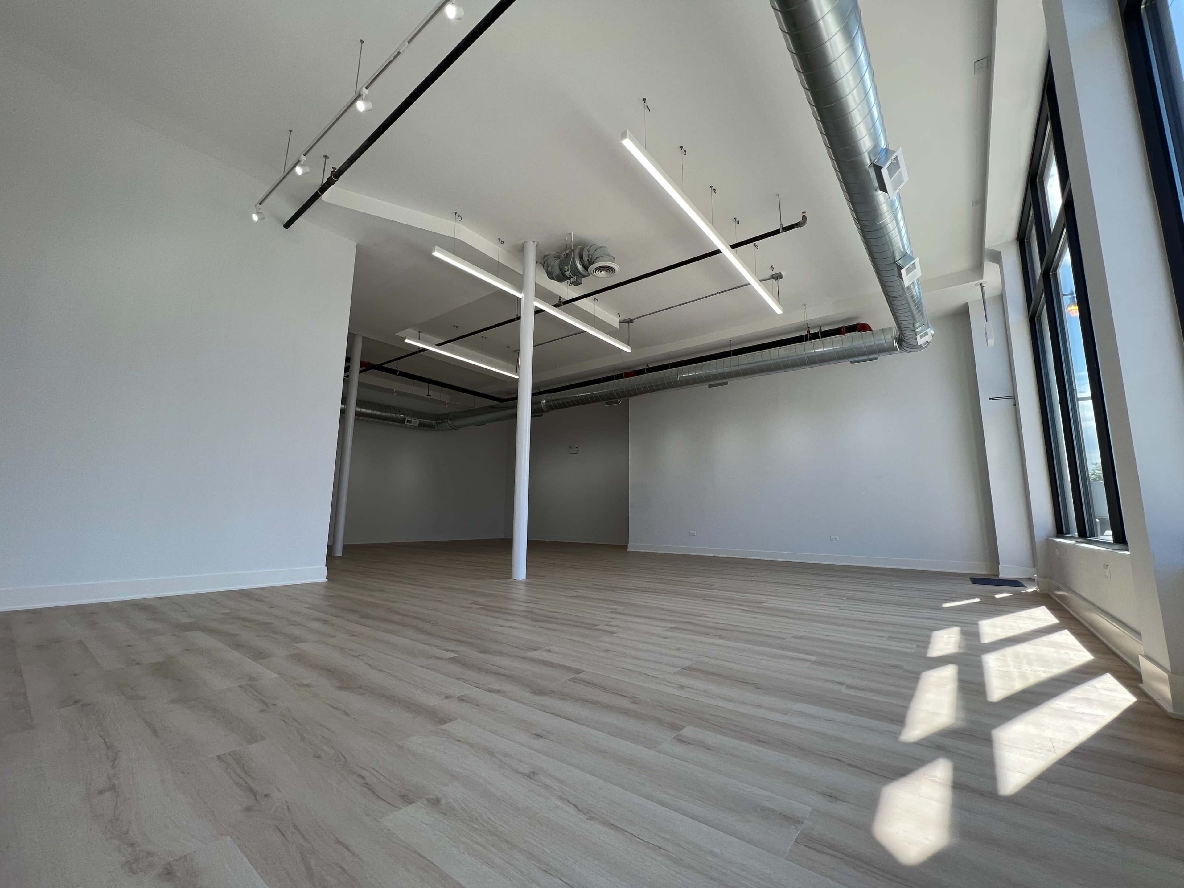 A spacious, unfurnished room with light-colored wooden flooring and large windows allowing natural light.