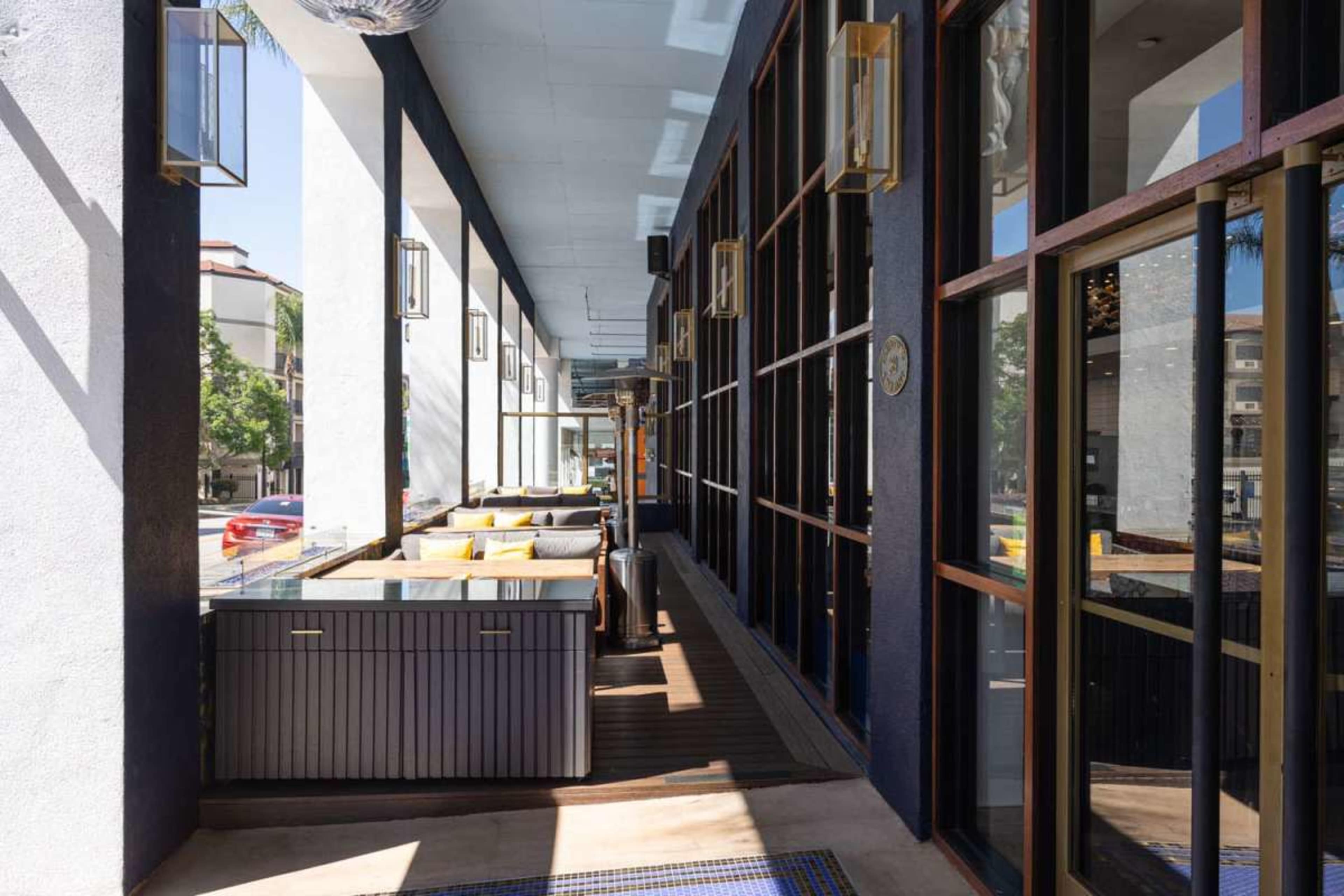 The image shows a modern outdoor seating area with large windows and a row of tables along a covered walkway.