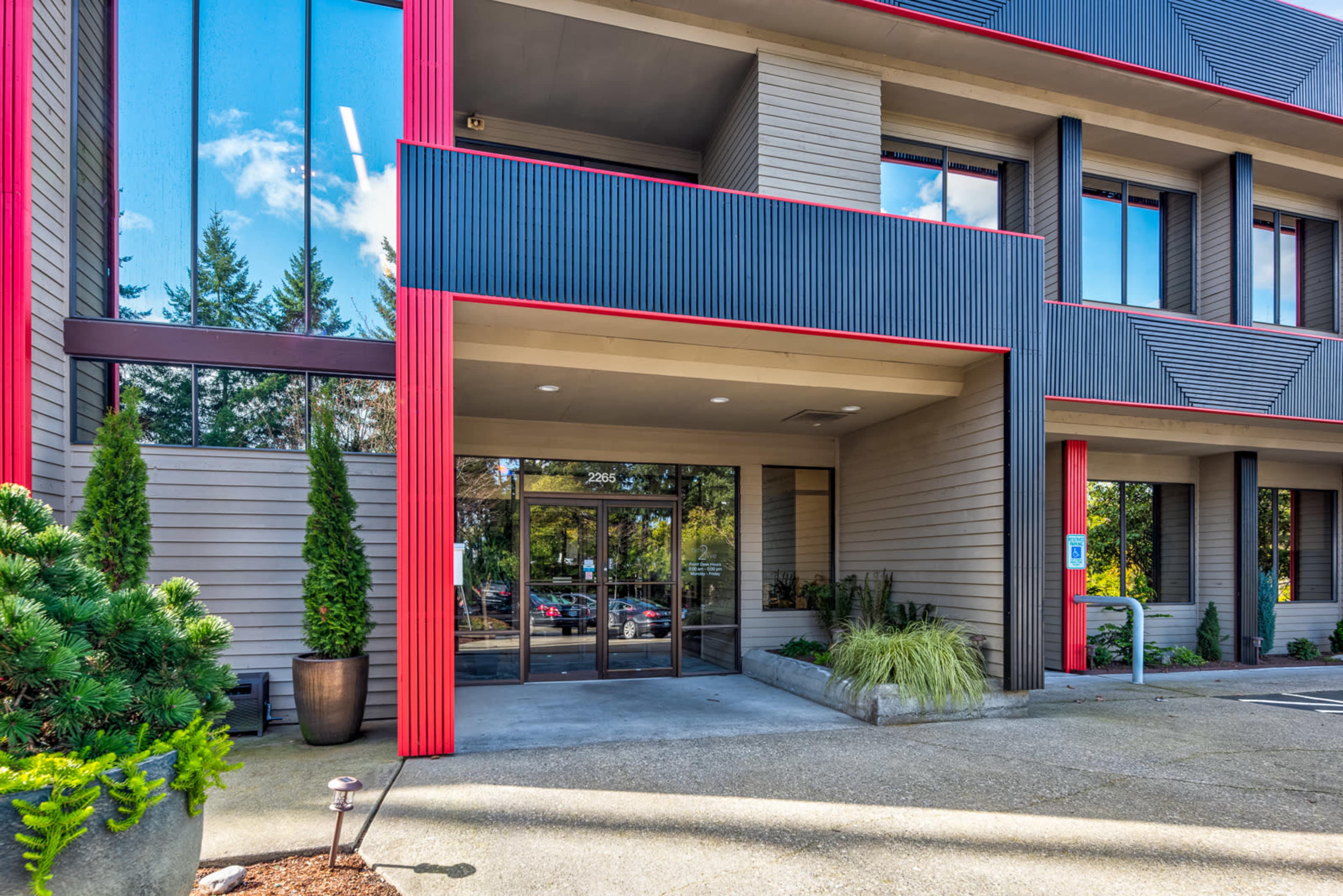 The entrance of a modern building with large glass doors and red accents framed by gray wood siding.