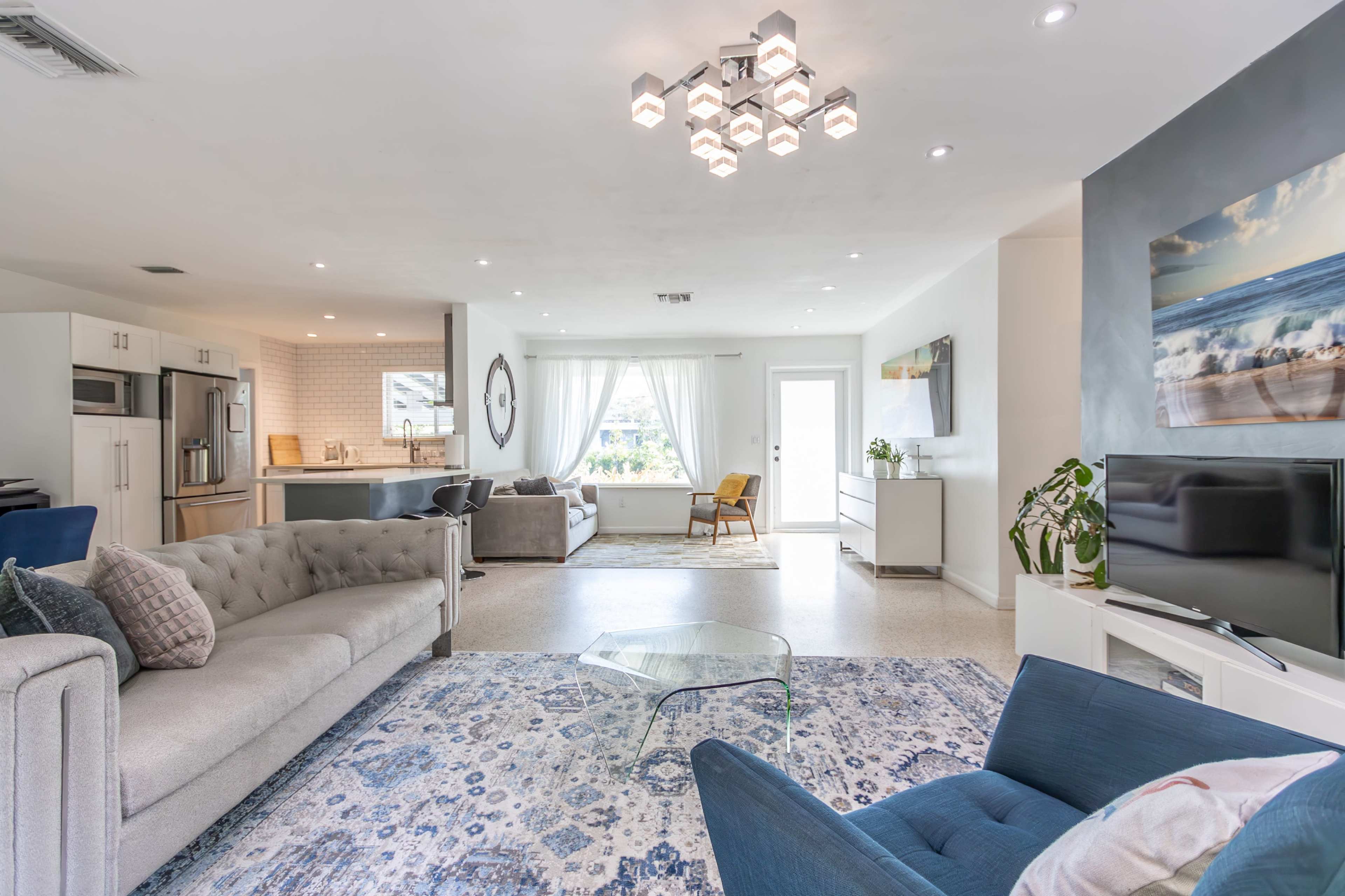The image shows a modern living room featuring a light-colored sofa, blue accent chairs, a glass coffee table, and a view of a kitchen area and outdoor space through a set of double doors.