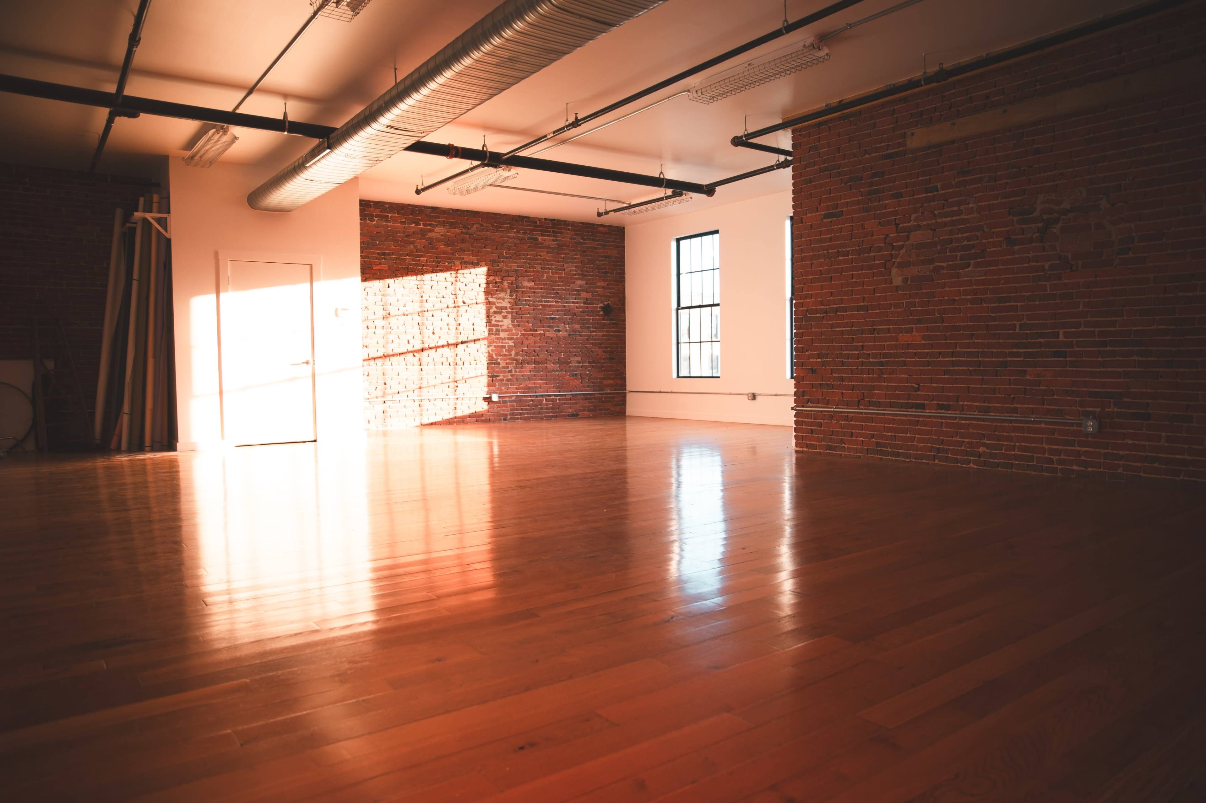The image shows a brightly lit, empty room with hardwood flooring and exposed brick walls.