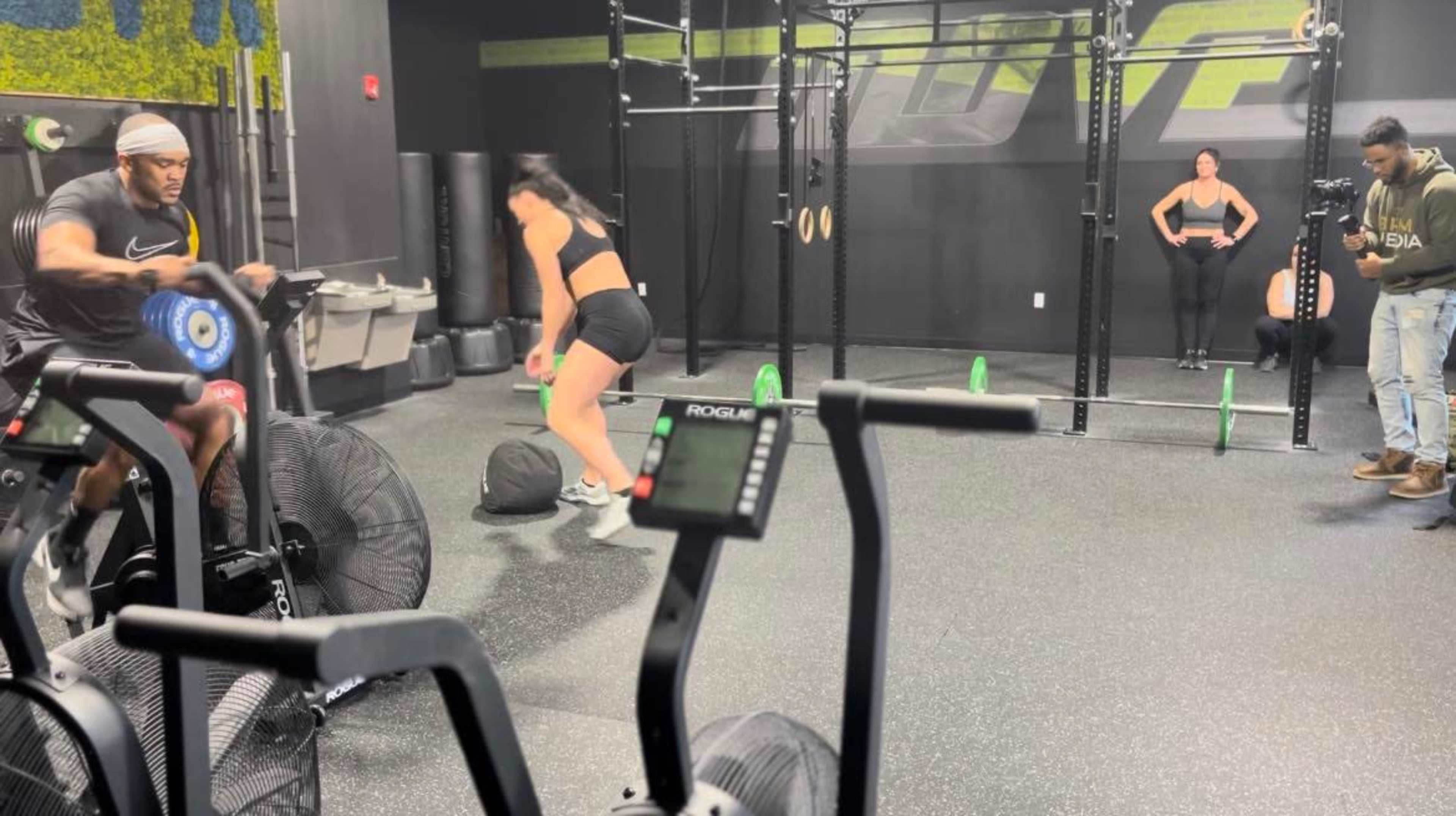 A man on an exercise bike and a woman performing a workout move are in a gym with multiple exercise equipment and a photographer capturing the scene.