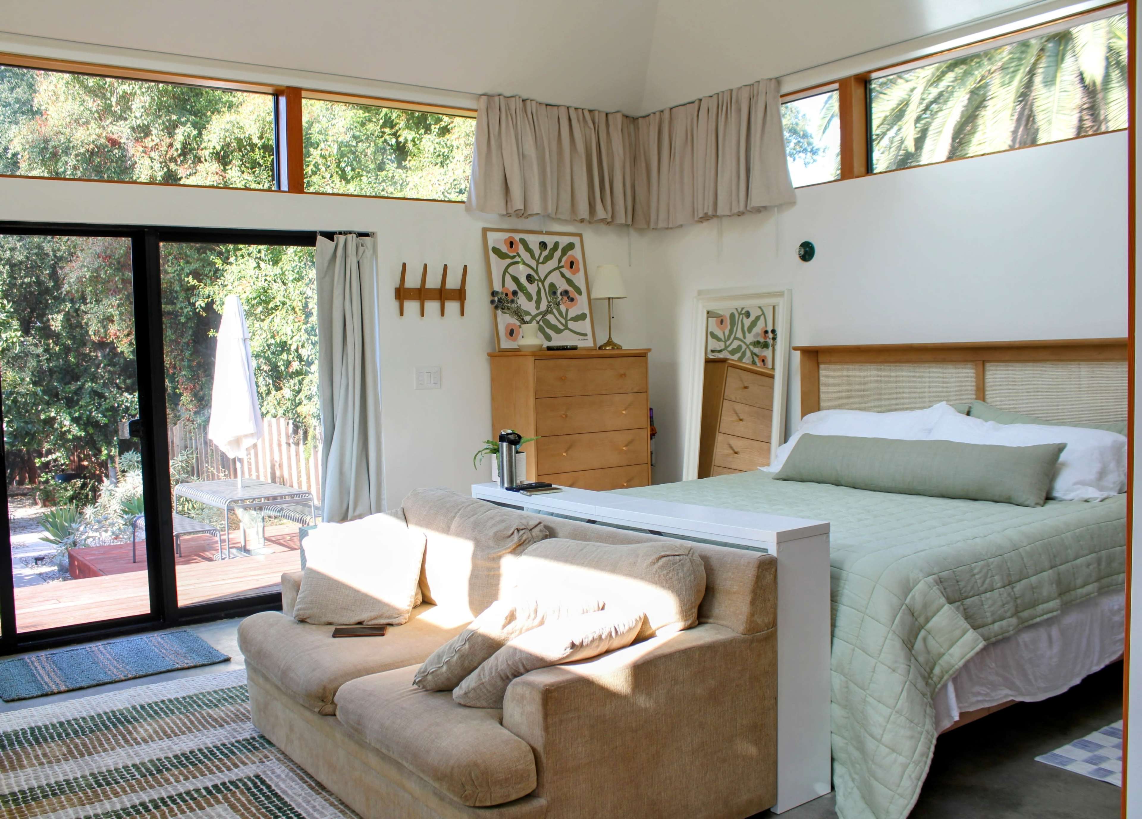 The image shows a bright, modern bedroom with a bed, a sofa, and sliding glass doors that lead to a deck outside.