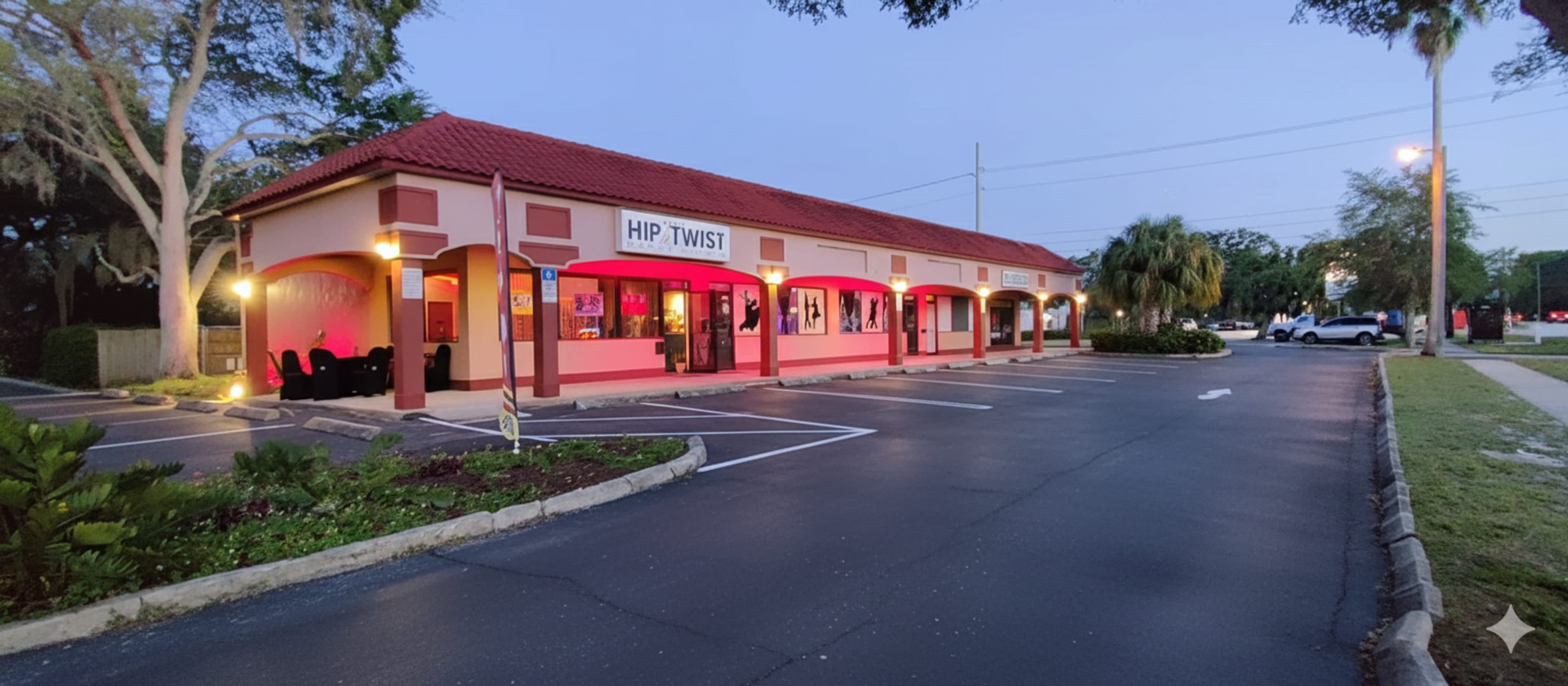 The image shows a single-story commercial building with a red-tiled roof and pink exterior, featuring several storefronts, including one labeled "HIP TWIST," set in a partially empty parking lot during twilight.