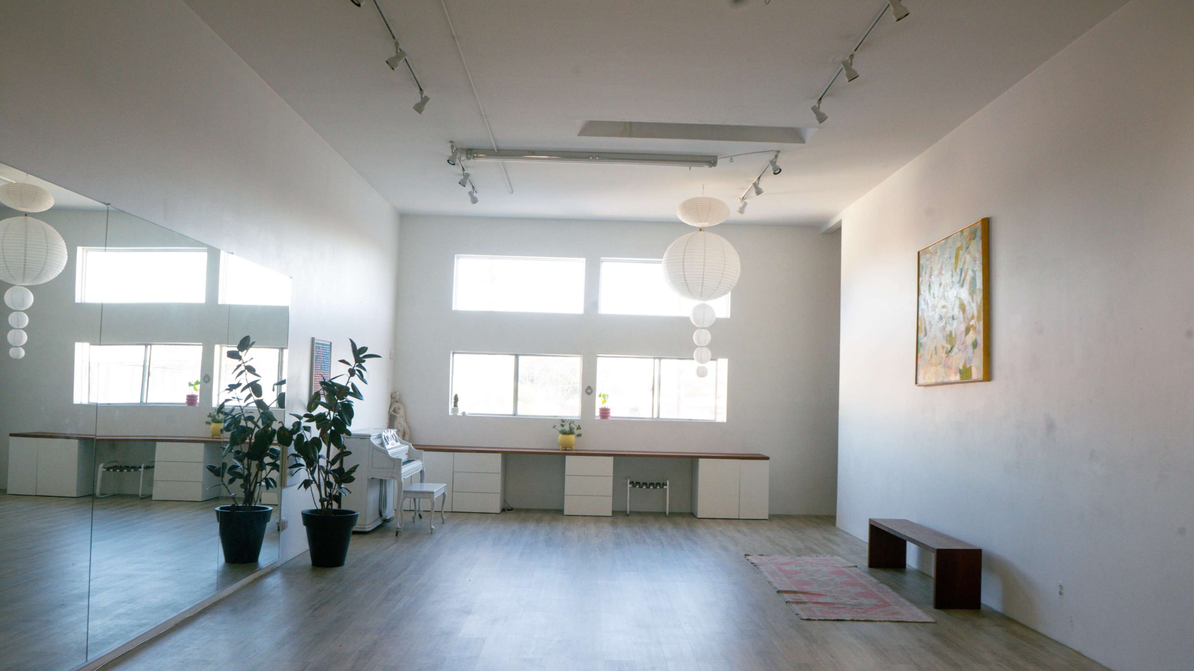 The image shows a spacious room with wooden flooring, large windows allowing natural light, a potted plant, a wooden bench, and minimal furnishings against a white wall.