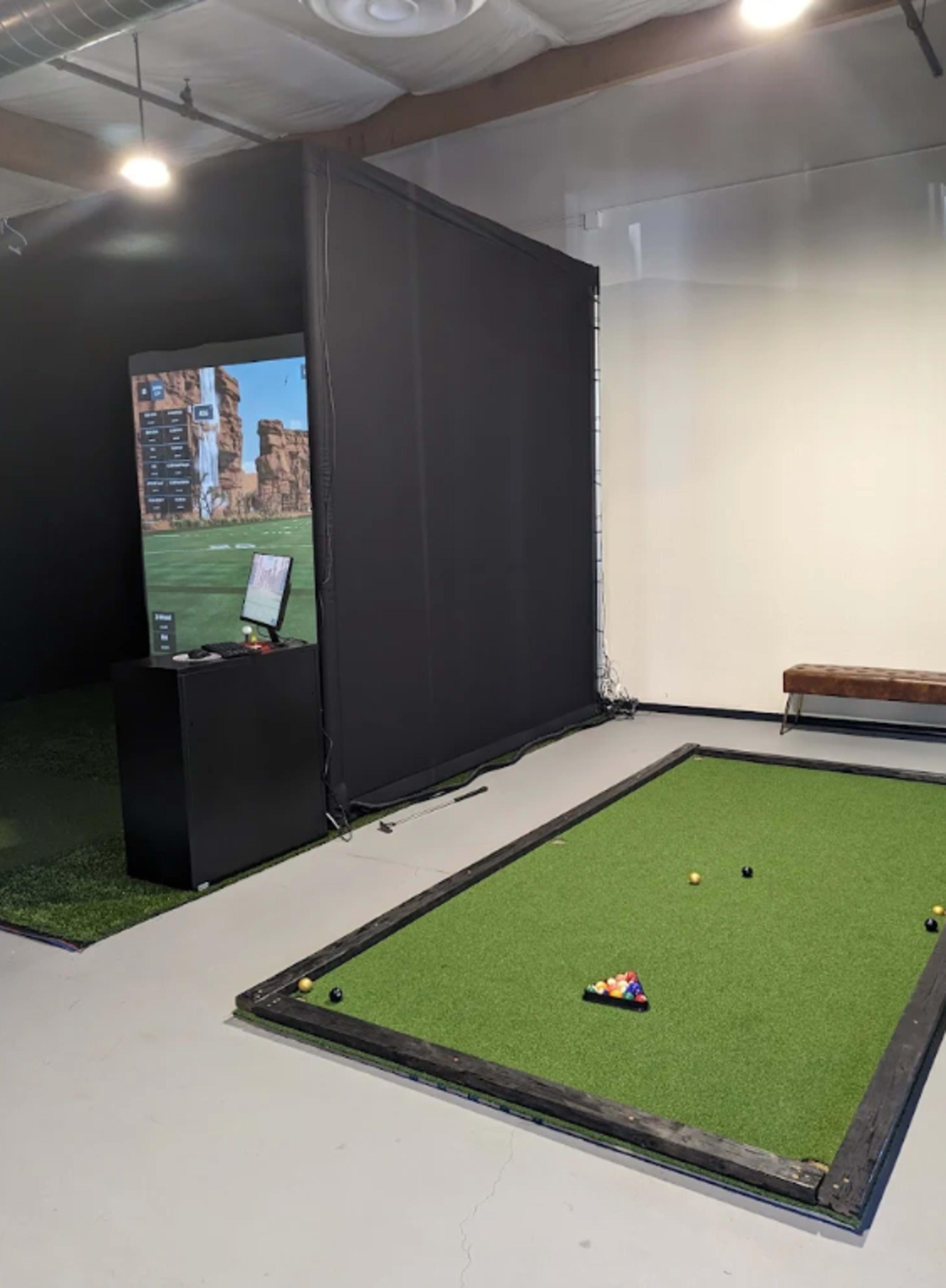 The image shows an indoor putting green area with a digital screen displaying a golf simulation, alongside a bench against a wall.