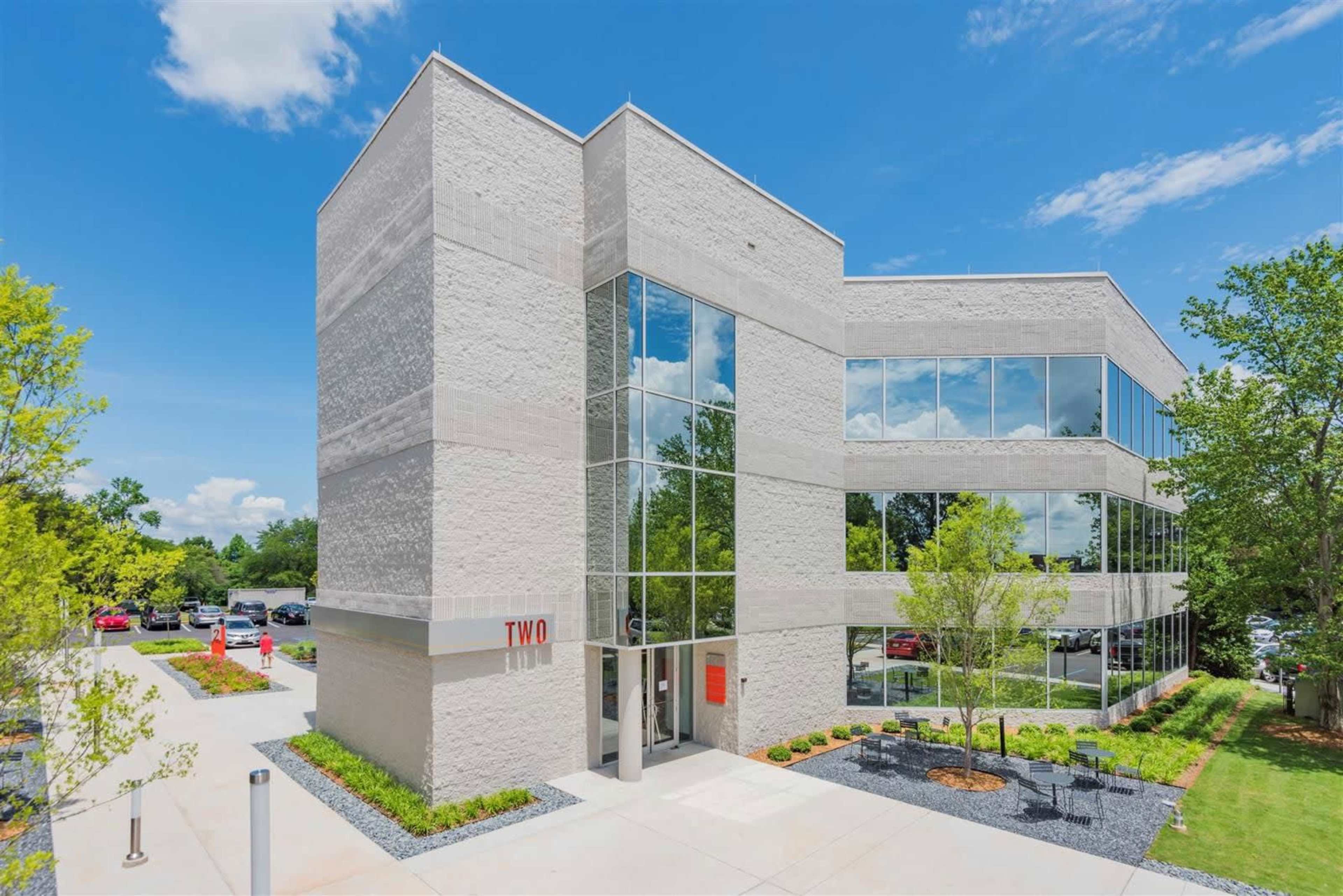 The image shows a modern, multi-story office building with large glass windows surrounded by landscaped greenery and a paved parking area.