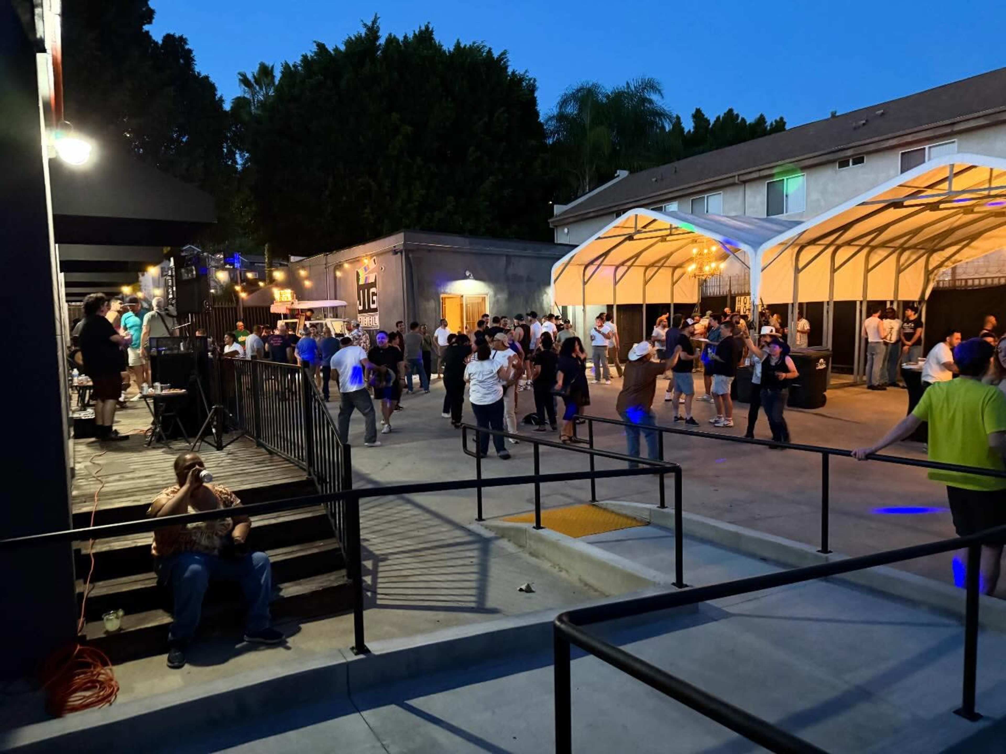 A crowd of people gathers at an outdoor event space, with some participants dancing and others socializing under a tented area as the evening light fades.