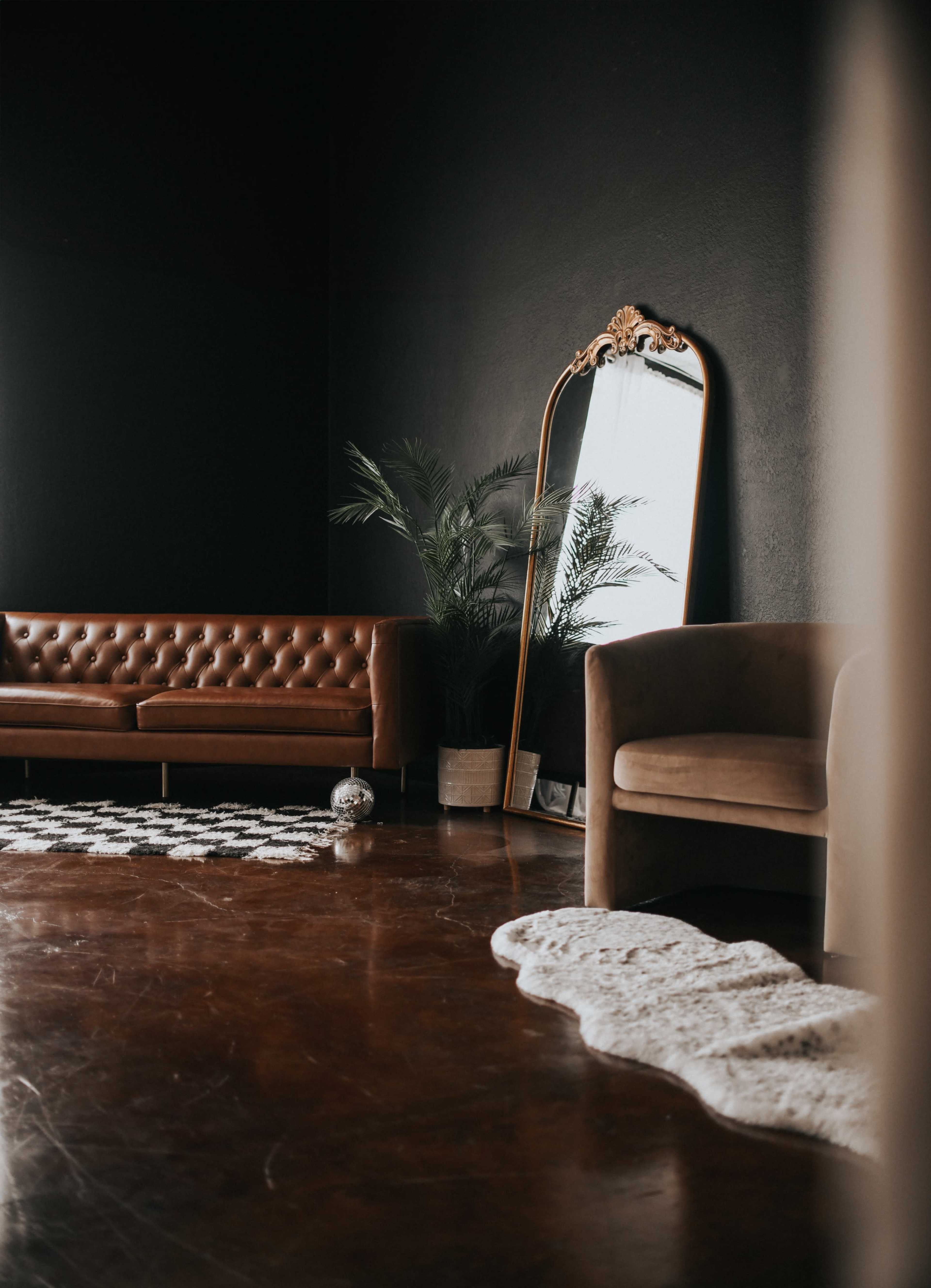 The image shows a modern living room featuring a brown leather sofa, a vintage-style mirror, a potted plant, and decorative rugs on a polished floor.