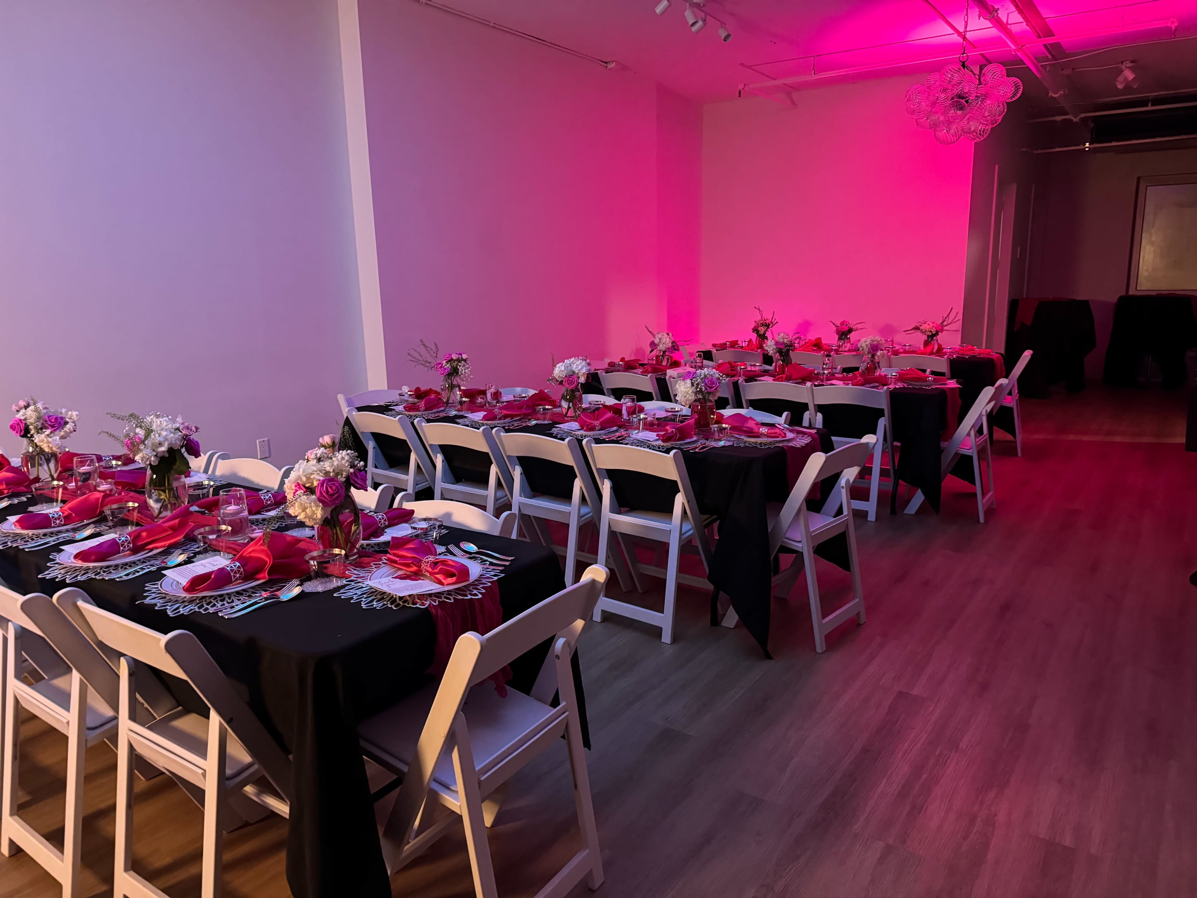 A banquet room is set up with tables draped in black and pink linens, adorned with floral centerpieces and matching place settings under pink lighting.