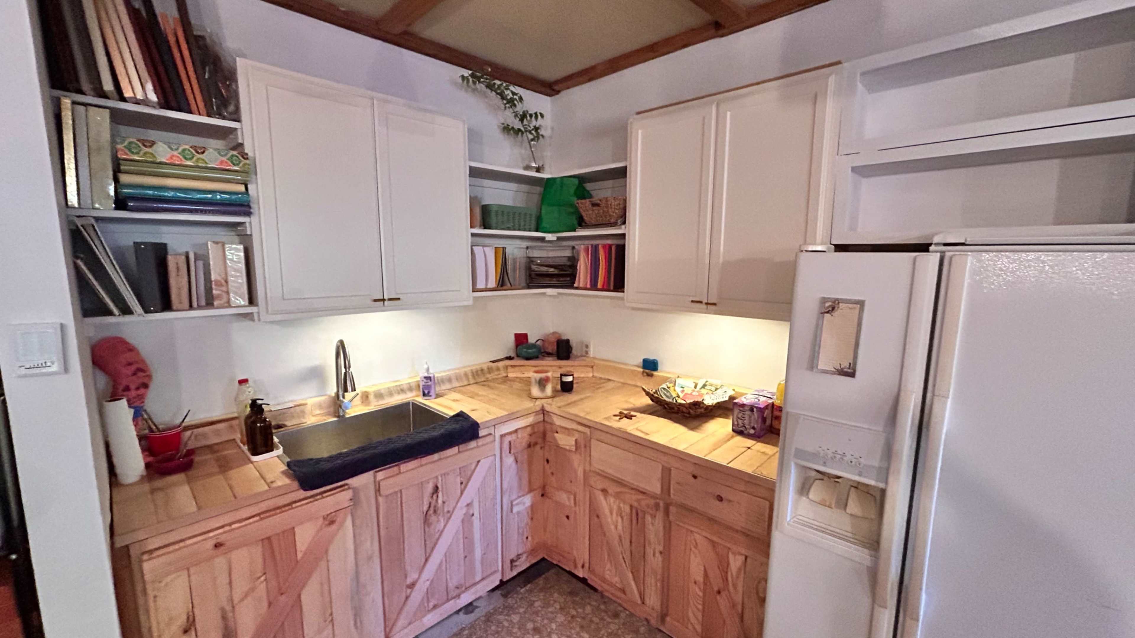 The image shows a small kitchen with wooden cabinetry, a stainless steel sink, and shelves filled with various items.