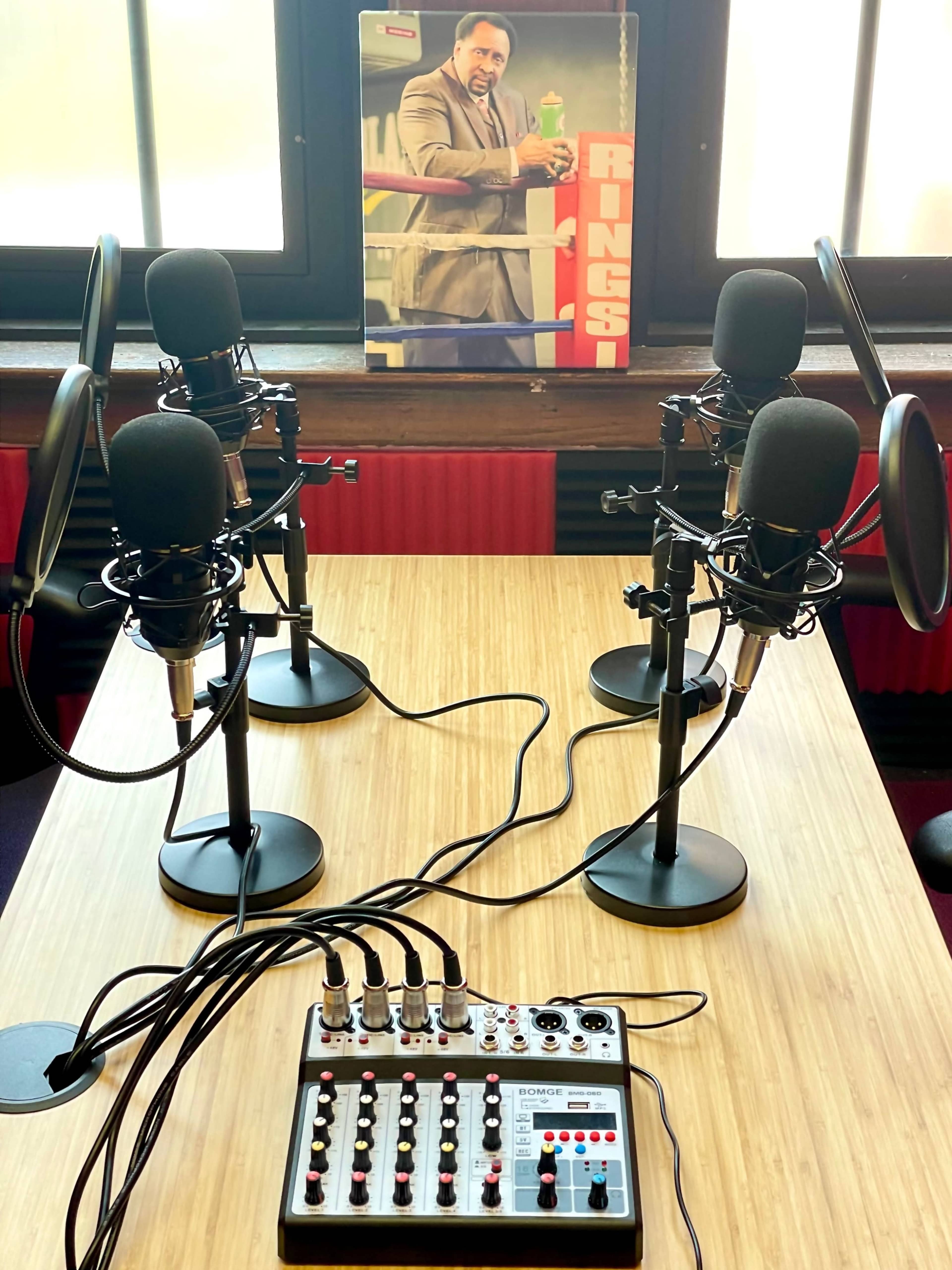 A podcast recording setup featuring four microphones, a sound mixer, and a poster of a man in a boxing ring in the background.