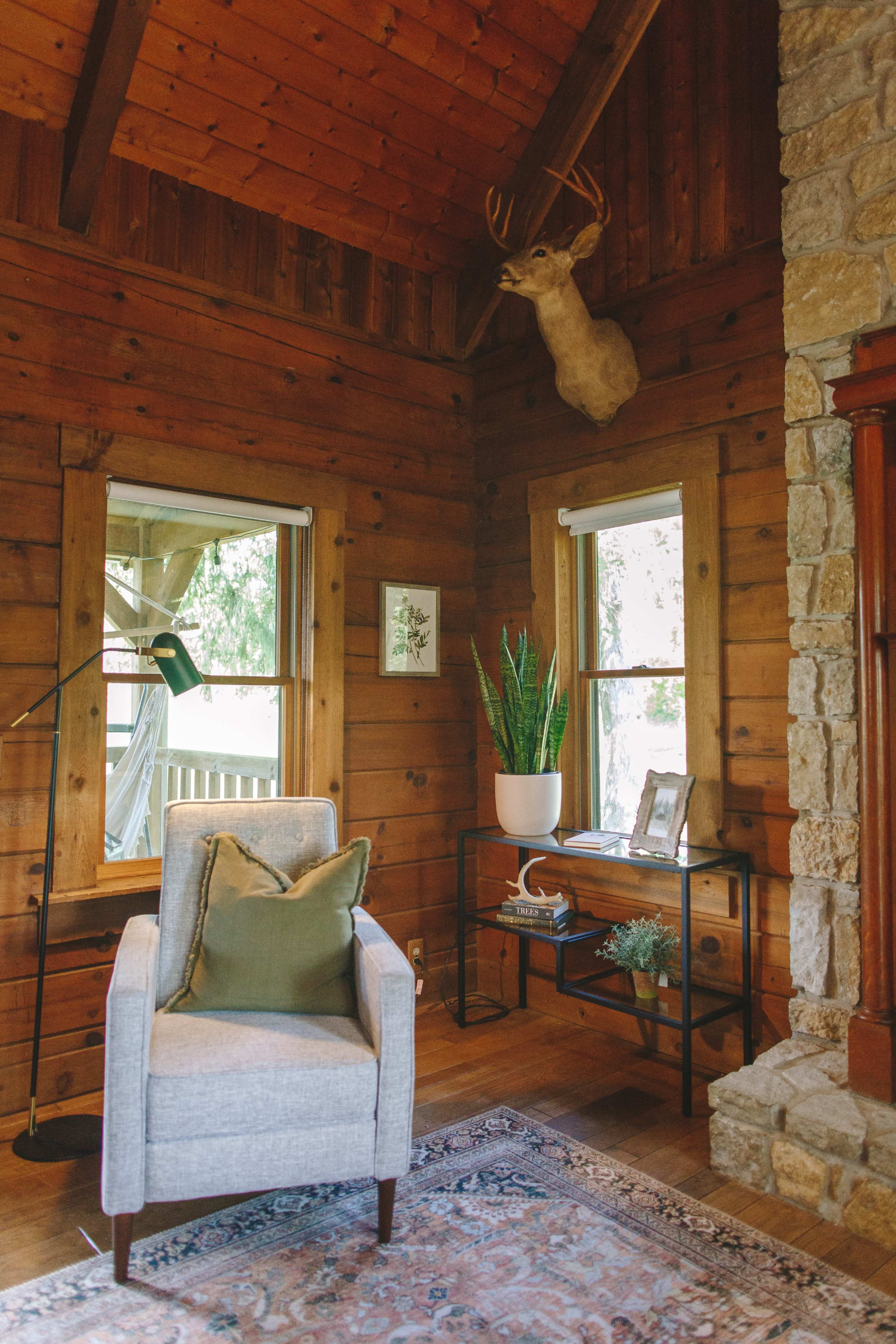 A cozy corner of a wooden room featuring an armchair, a mounted deer head, a lamp, and a small console table with plants and decor items.