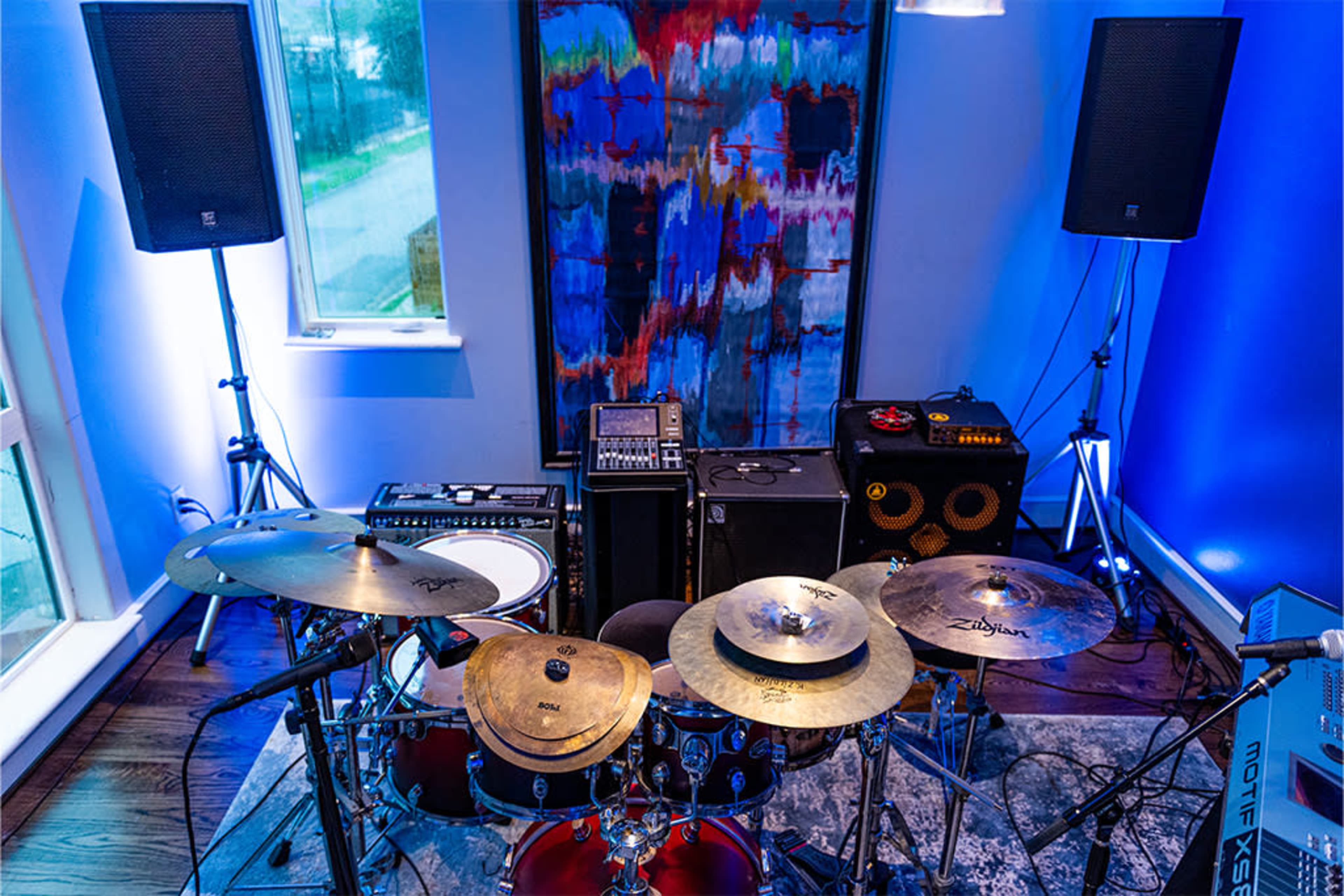 A drum set positioned in a well-lit room with speakers, an amplifier, and abstract artwork on the wall.