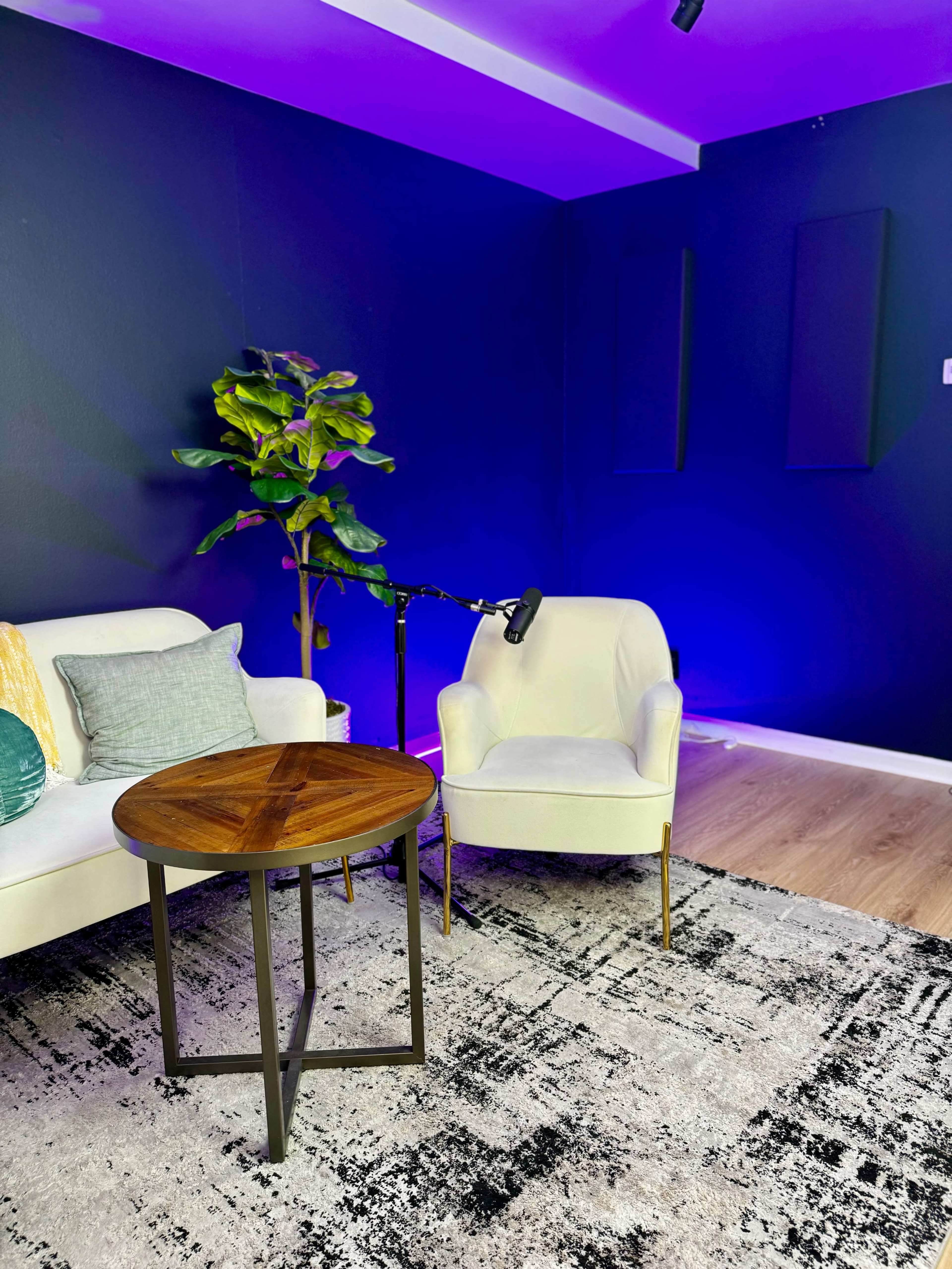 A cozy seating area features a white sofa and an armchair beside a wooden table, with a plant and a microphone on a stand in a room illuminated by blue lighting.