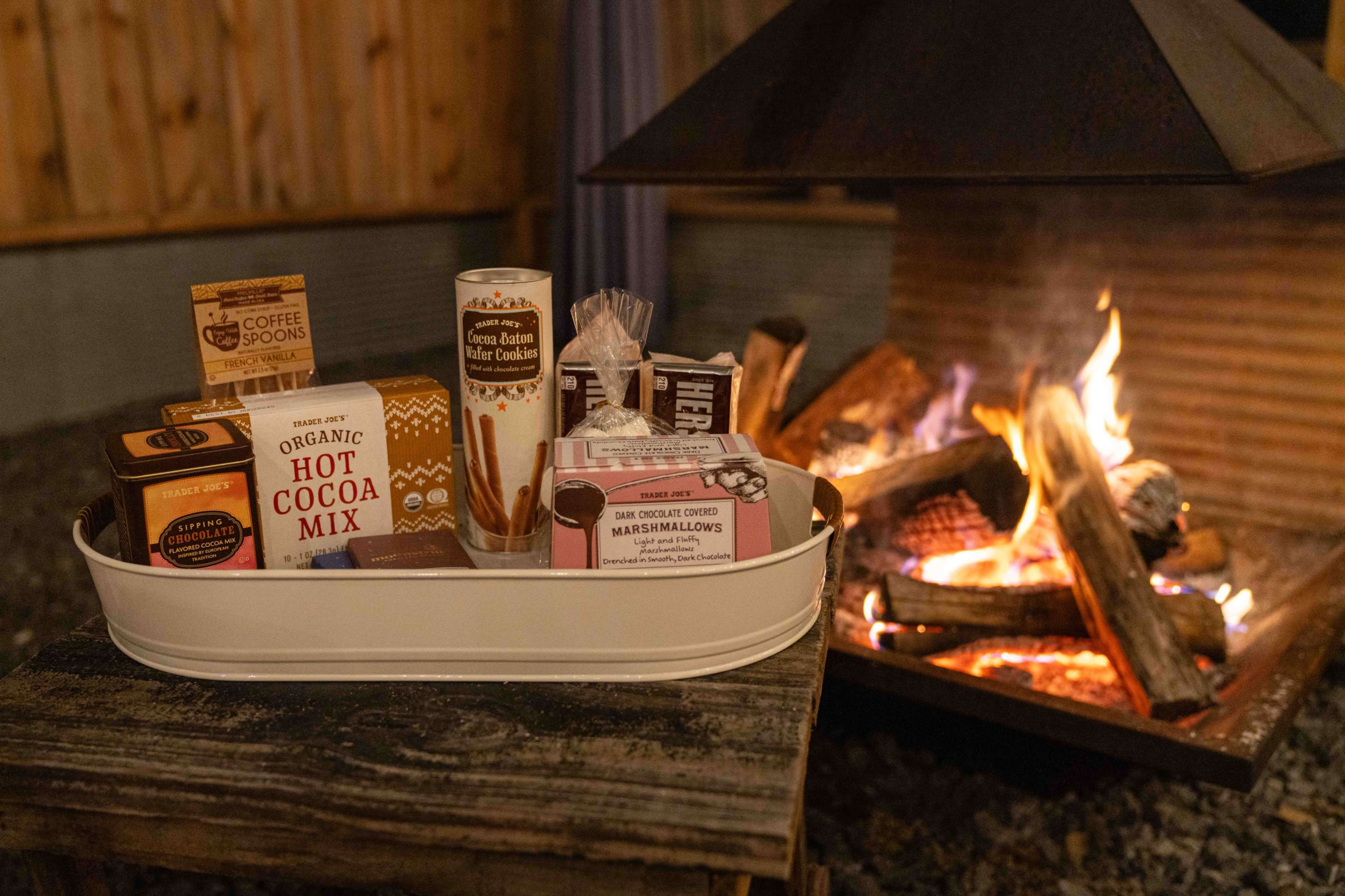 A tray filled with hot cocoa mix, marshmallows, and cookies is placed beside a crackling fire in a rustic outdoor setting.