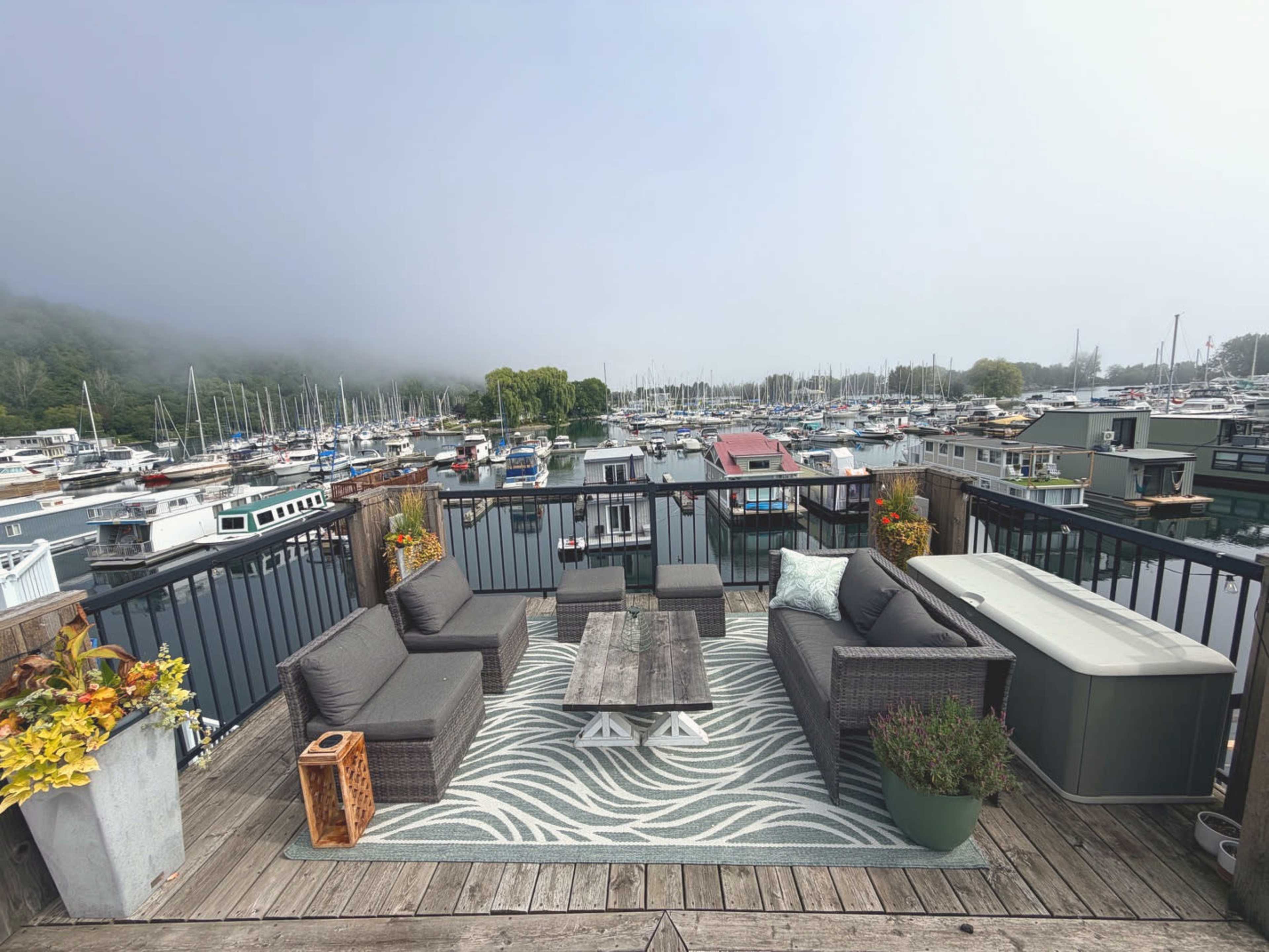 The image shows a spacious deck with outdoor furniture overlooking a marina filled with boats, set against a backdrop of fog-covered hills.