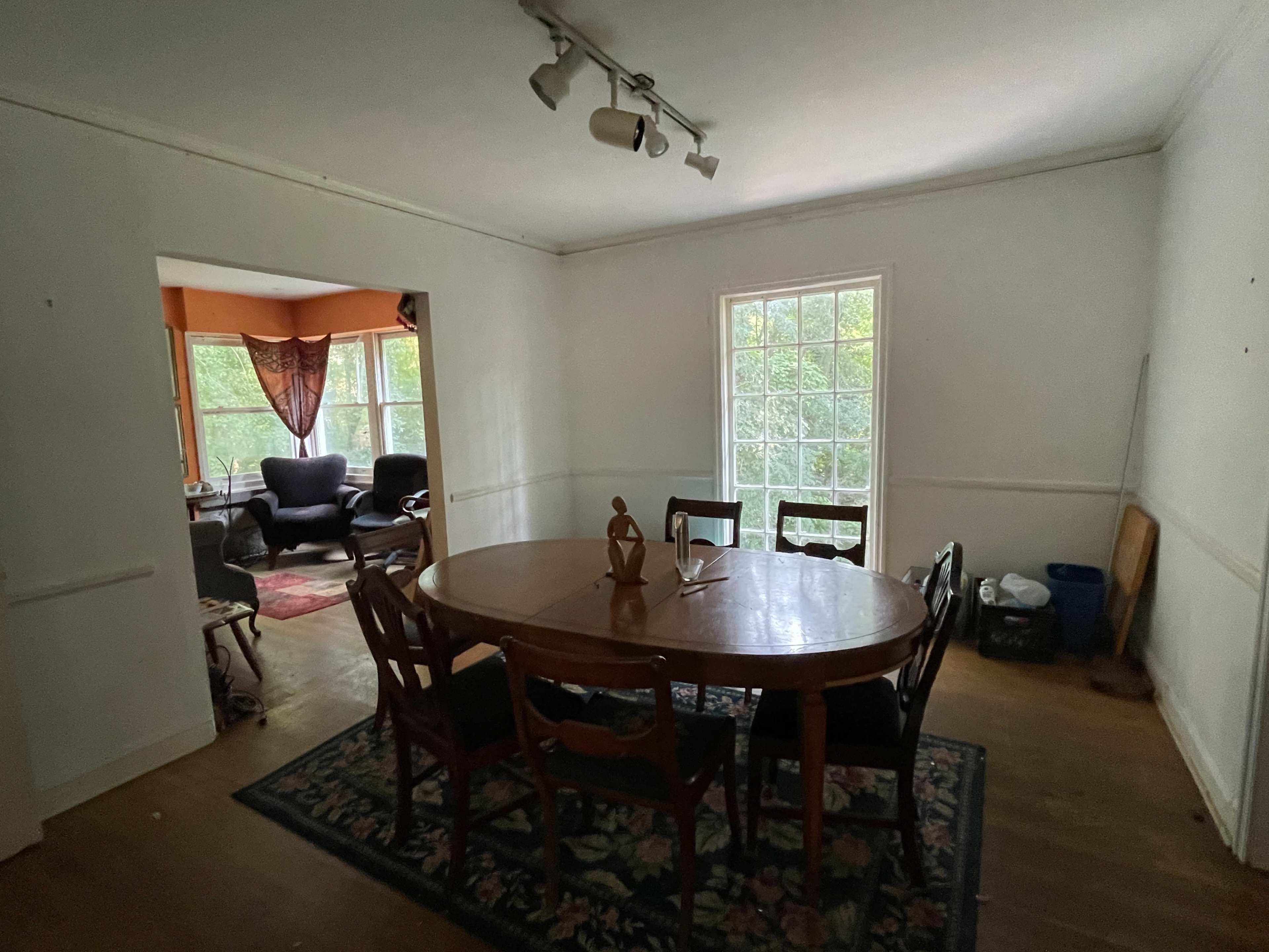 The image shows a dining area with a round wooden table surrounded by chairs, adjacent to a room with large windows and a seating area.