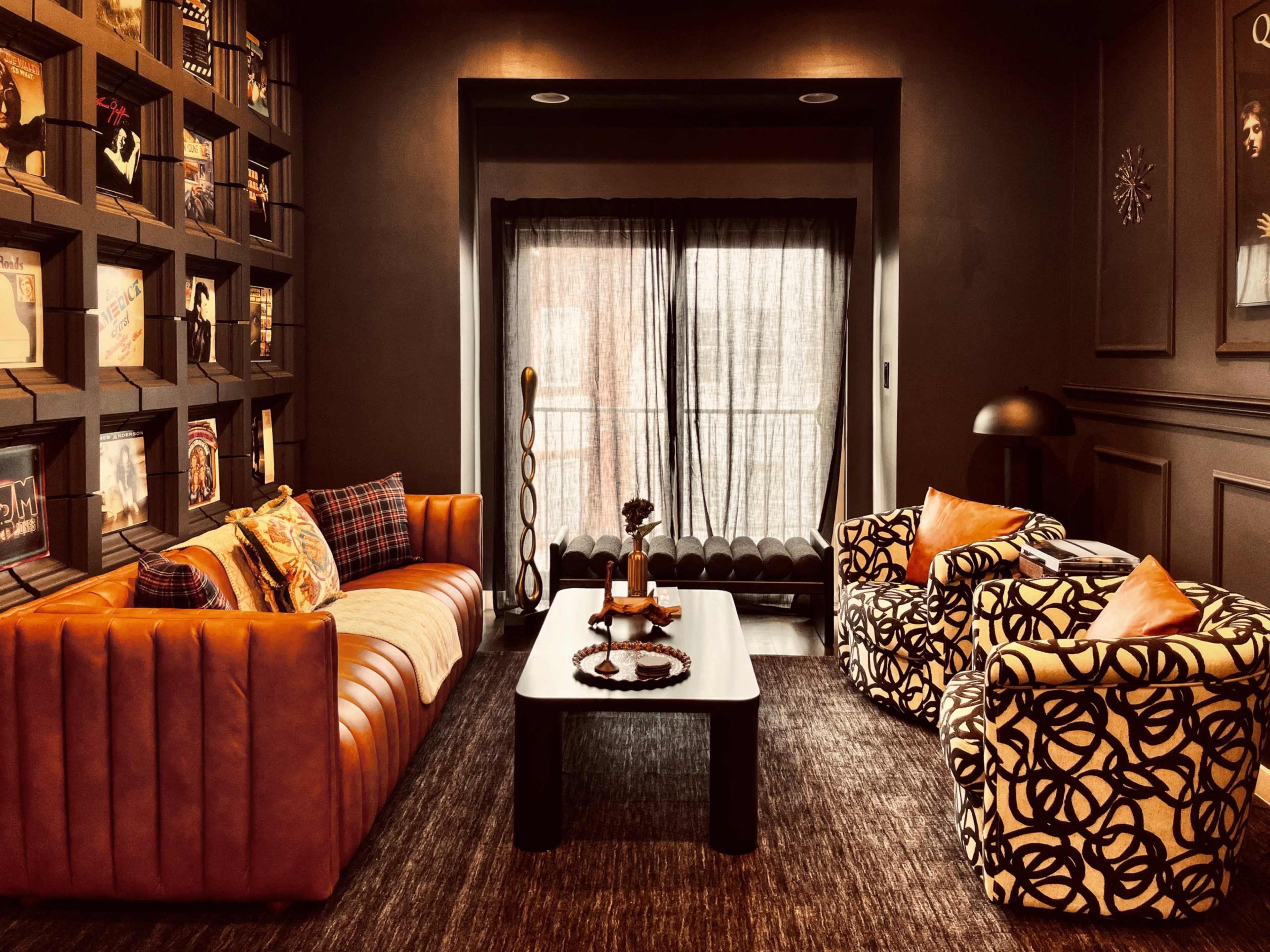 A stylish living room with a brown leather sofa, patterned armchairs, a dark coffee table, and a large window draped with sheer curtains.