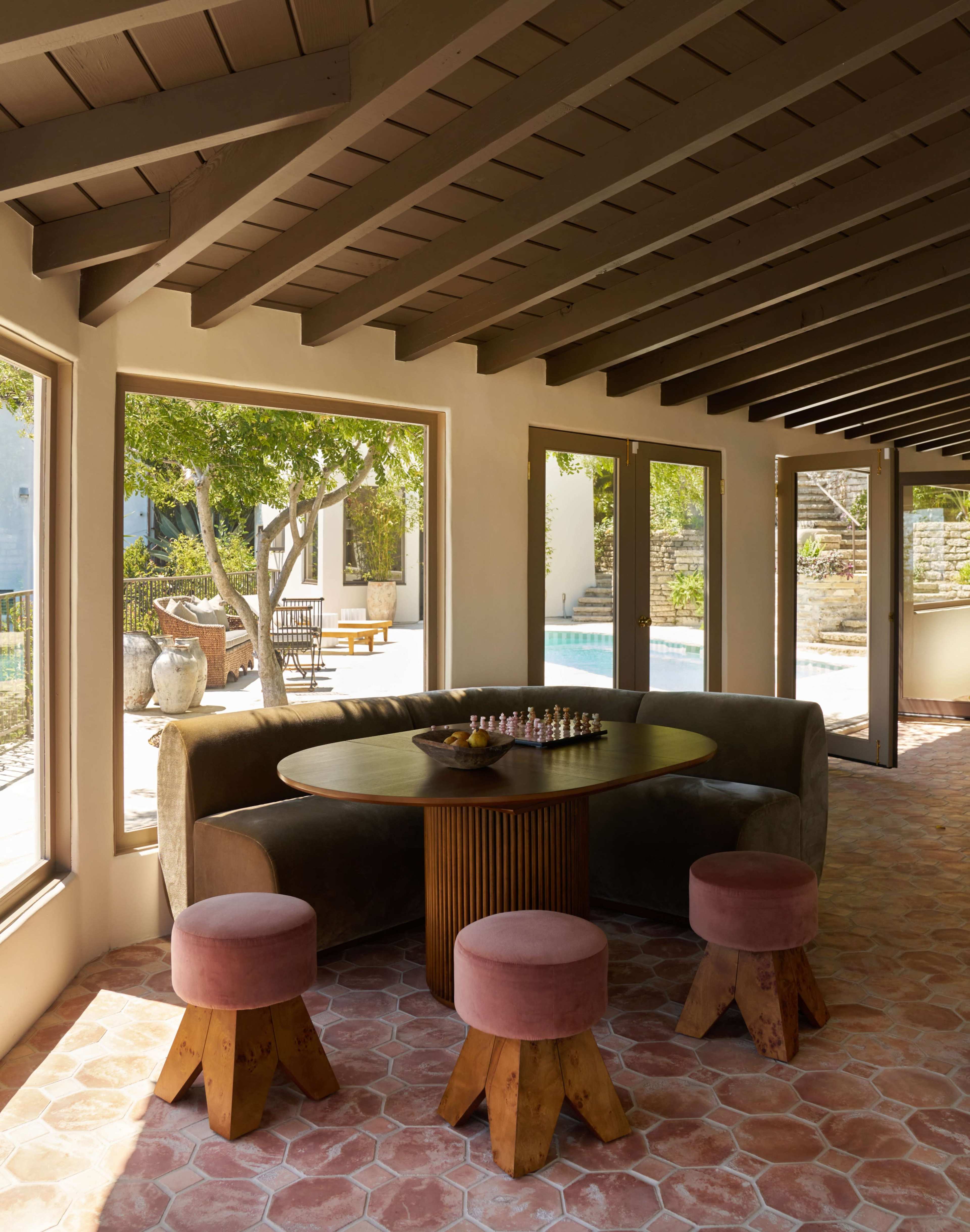 The image shows a modern indoor seating area featuring a round table with a chess set, surrounded by upholstered stools, and large windows overlooking a pool area.