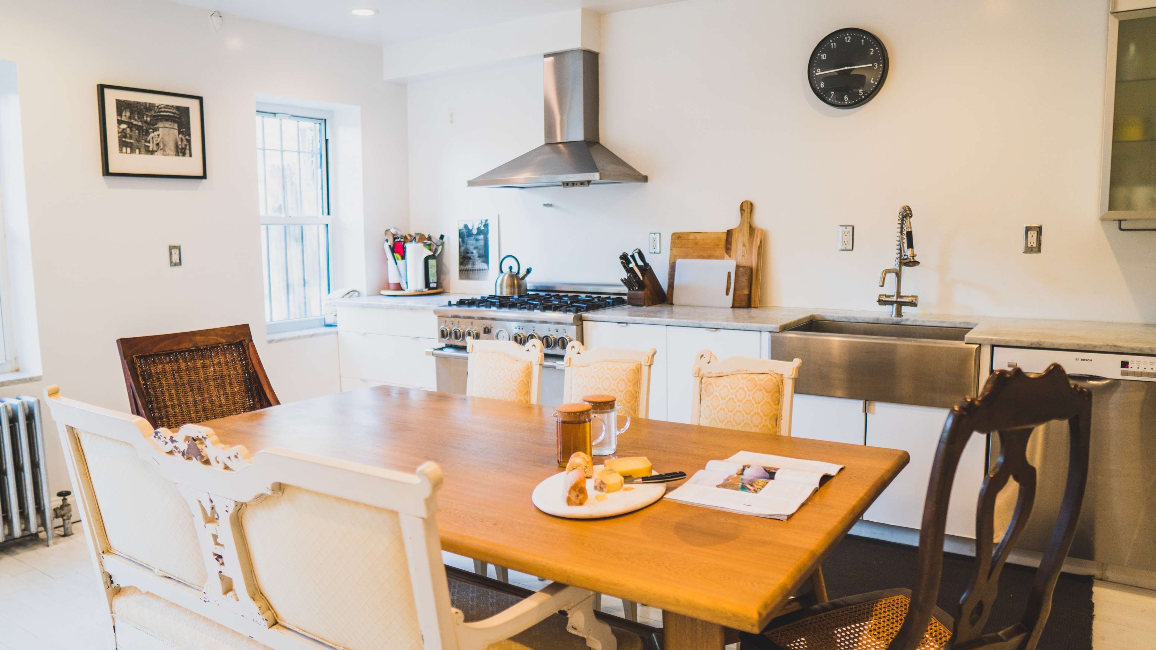 A modern kitchen features a wooden dining table with chairs, a stainless steel sink and stove, and wall-mounted cabinets.