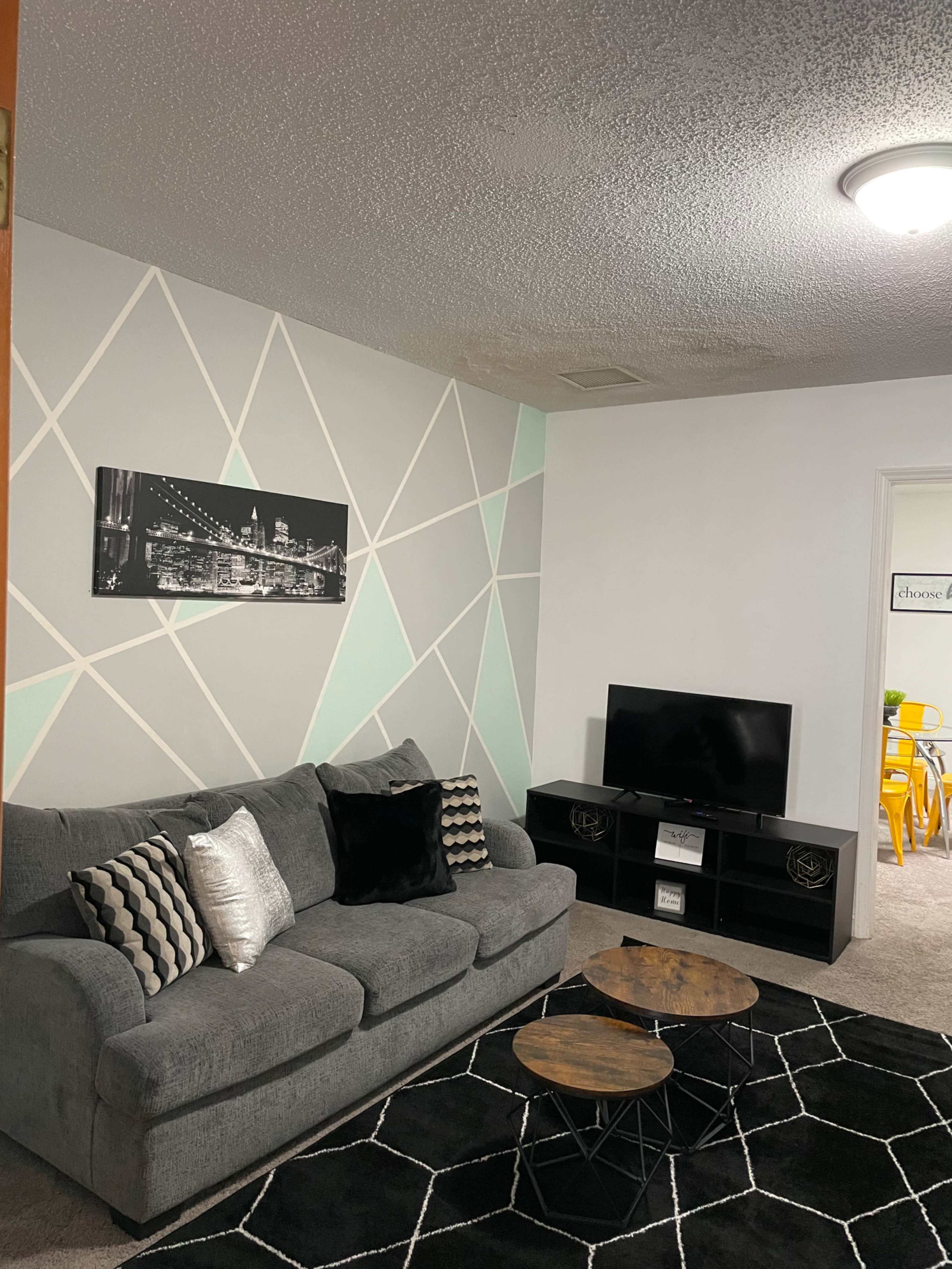 A living room featuring a gray sofa with decorative pillows, two wooden coffee tables, a black television stand, and a geometric patterned accent wall.