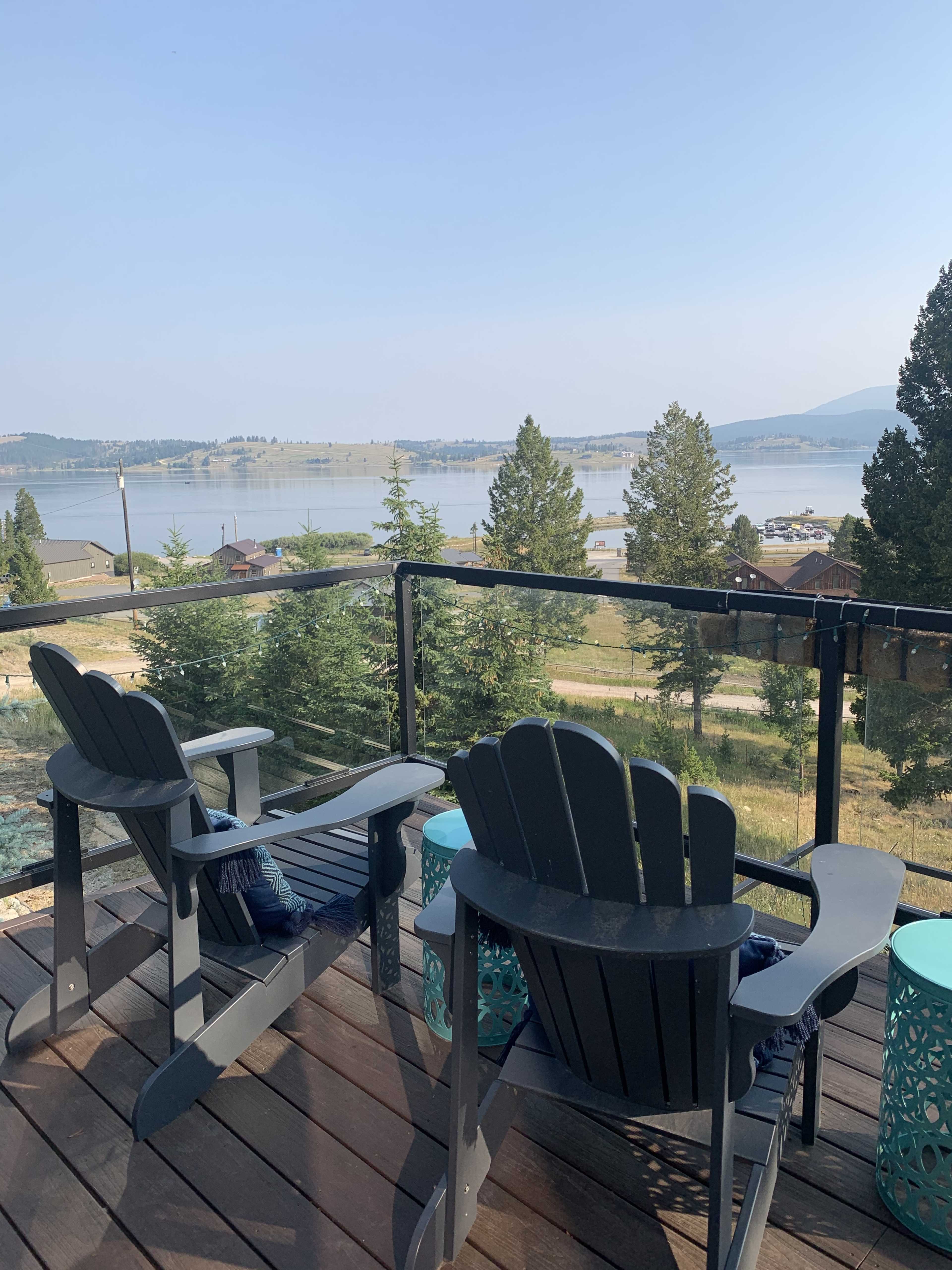 Two wooden Adirondack chairs are placed on a deck overlooking a lake with trees and distant hills in the background.