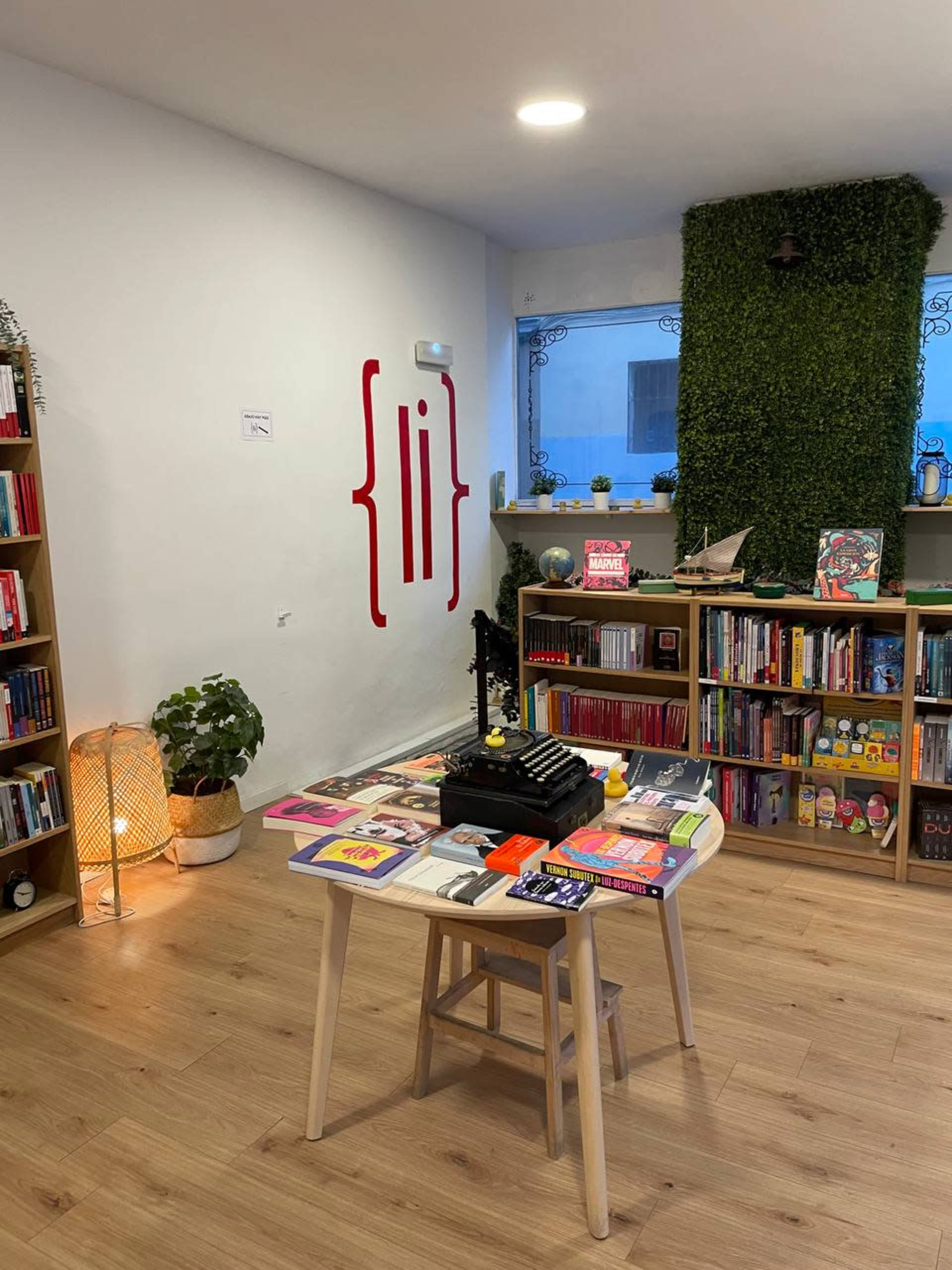 The image shows a cozy reading space with bookshelves filled with various books, a small table displaying books and a decorative object, and a wall featuring minimalist red letters.