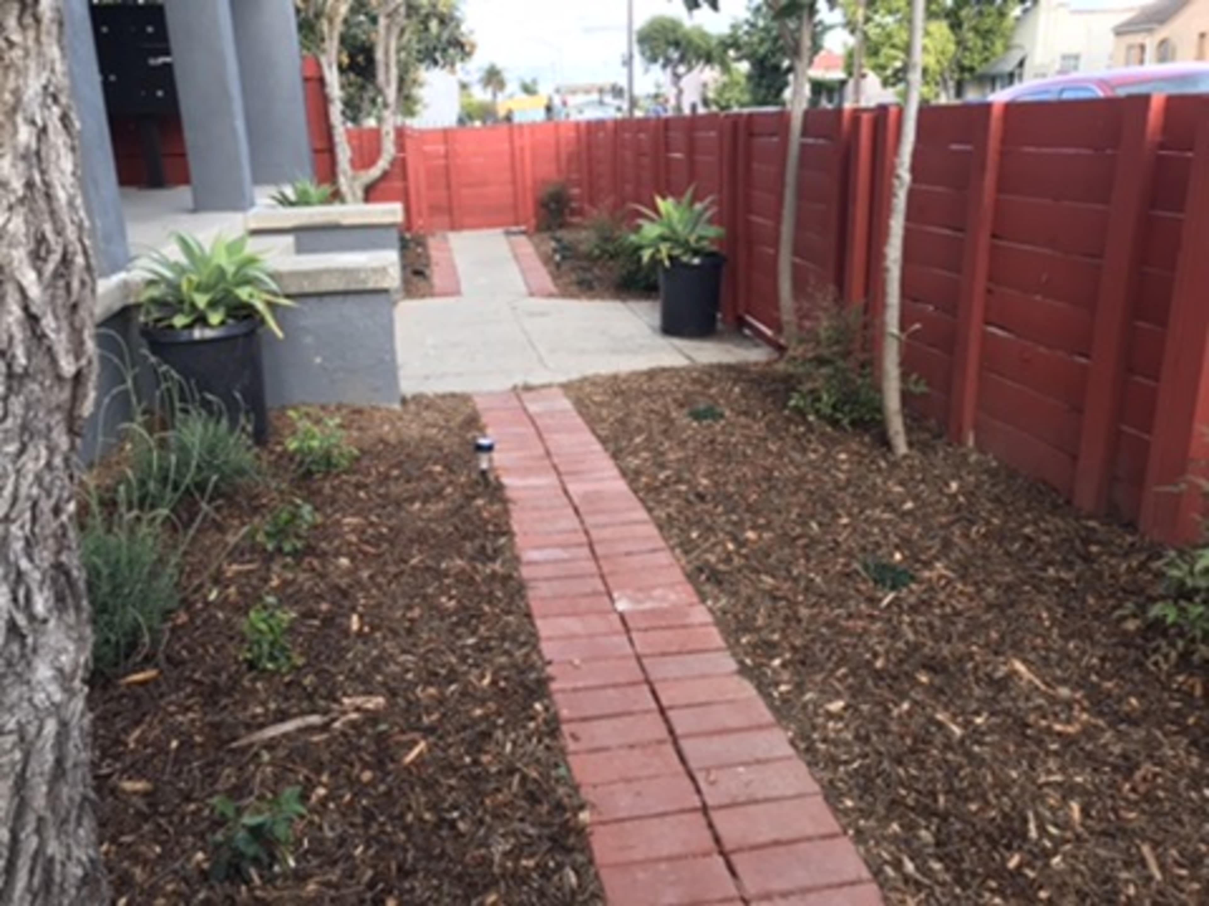 A pathway made of brick surrounded by landscaped areas with mulch and potted plants, leading past a red wooden fence.